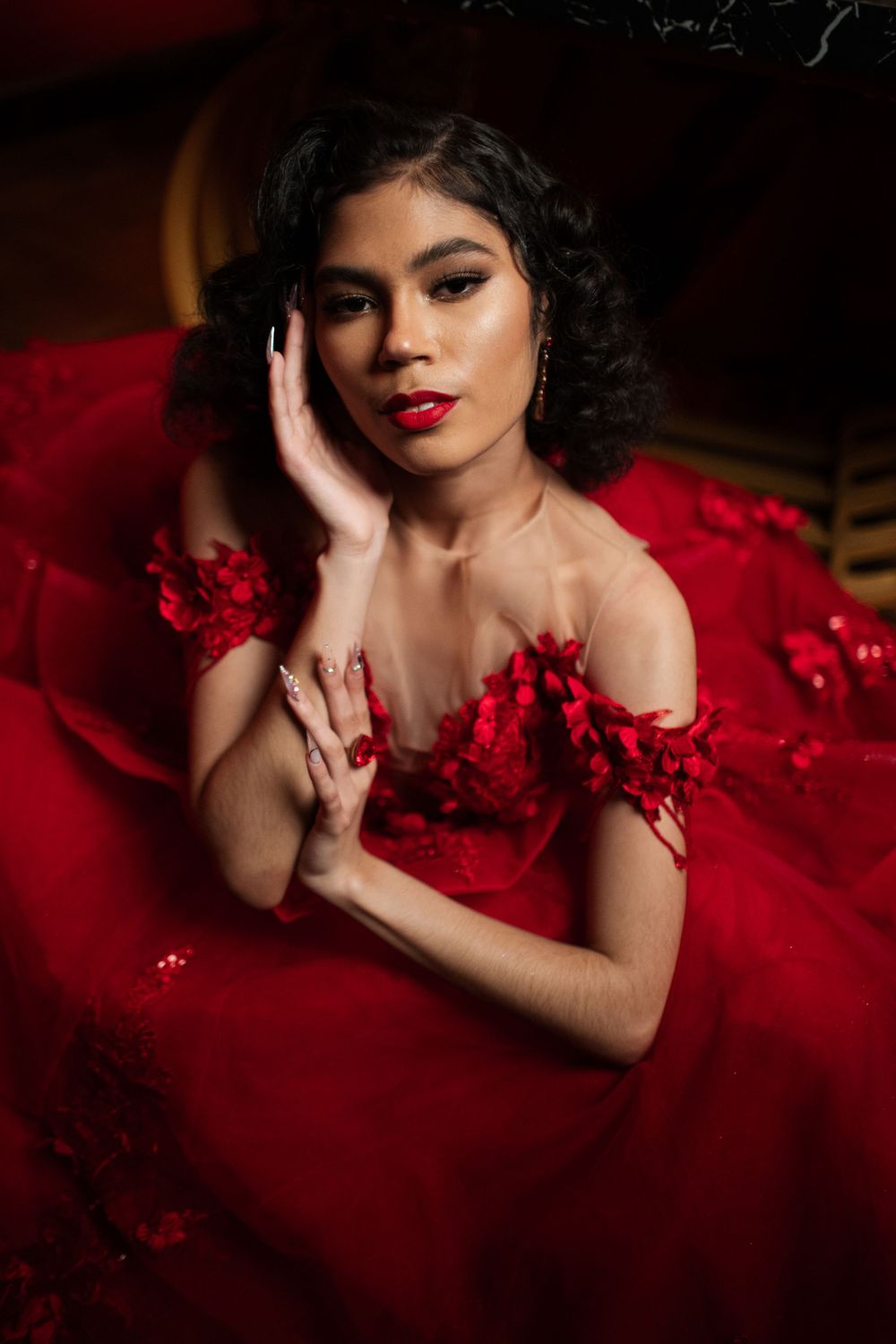 Dramatic portrait of model in luxurious red dress with red flowers against dark background.