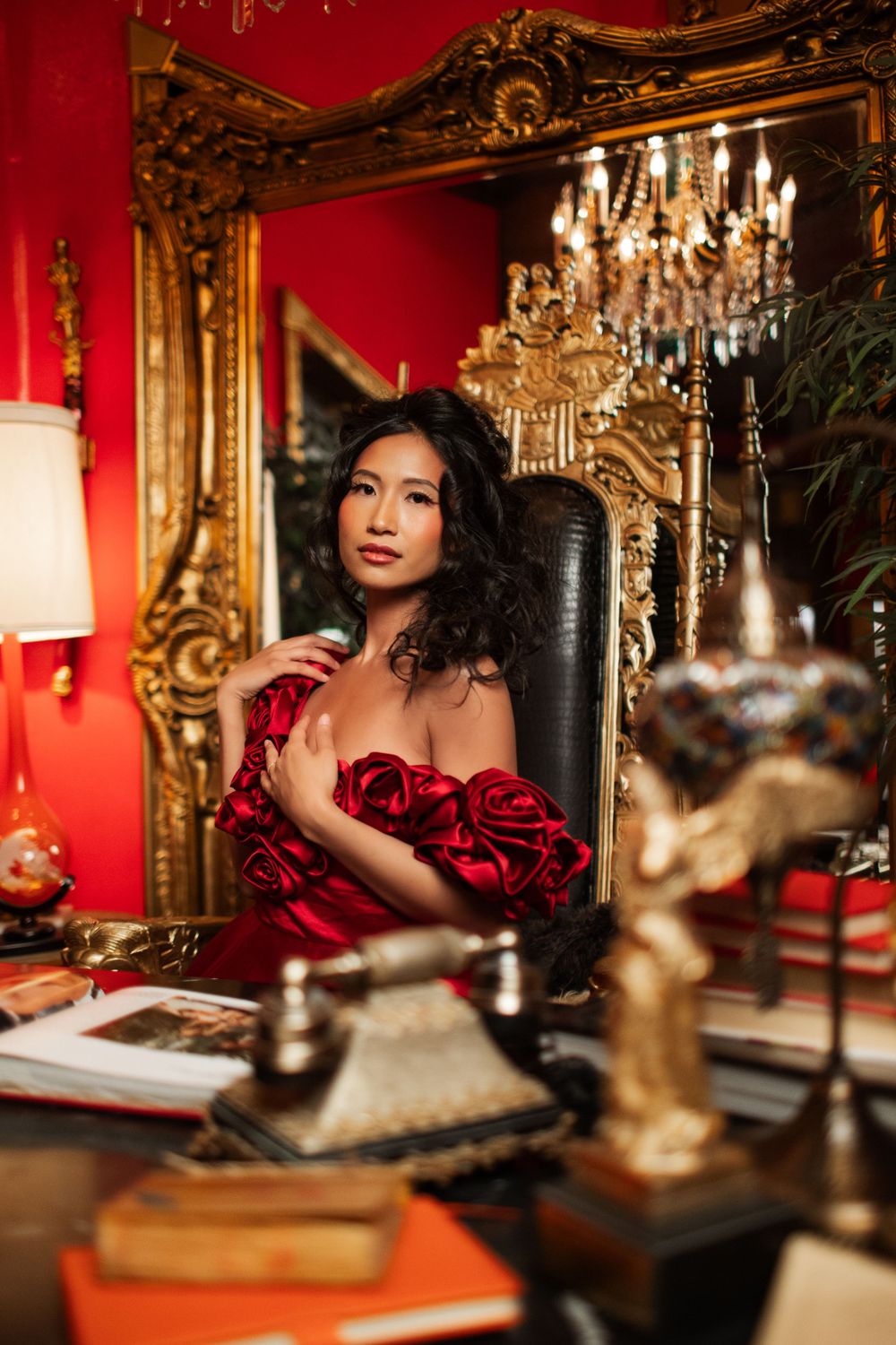 Elegant portrait in luxurious red room with ornate gold mirror, crystal chandelier and antique desk setting.