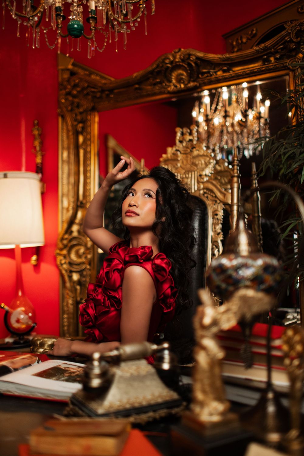 Glamorous woman in red floral dress poses thoughtfully in luxurious room with ornate gold mirror and crystal chandeliers.