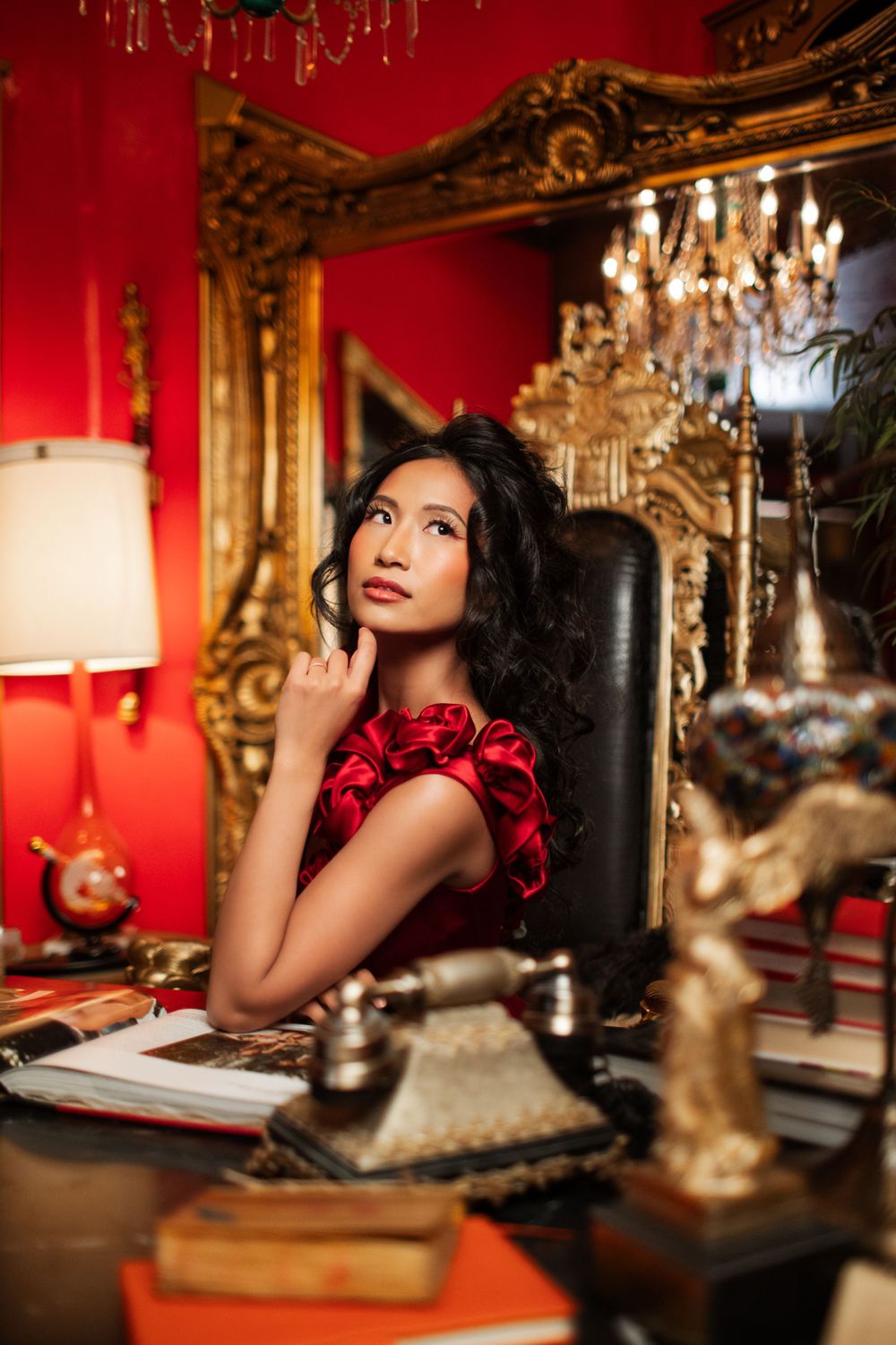 Fashion portrait in luxurious red room with ornate gold mirror and chandelier featuring red ruffled evening dress.