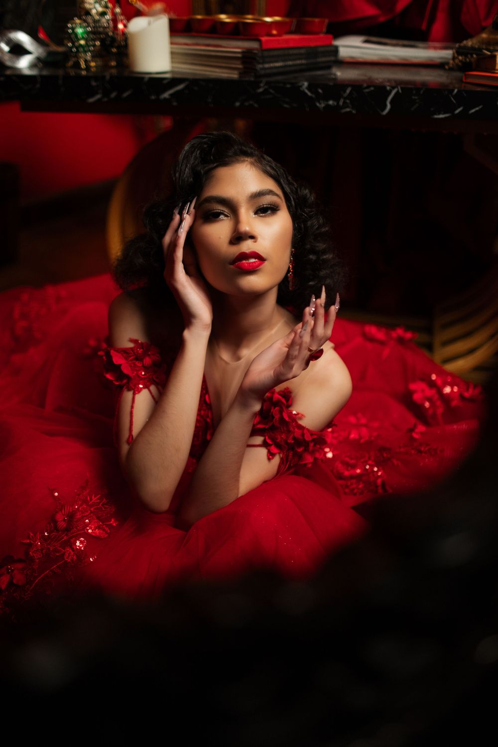 Artistic portrait in a deep red setting featuring dramatic lighting and a flowing crimson dress with festive decorative elements.