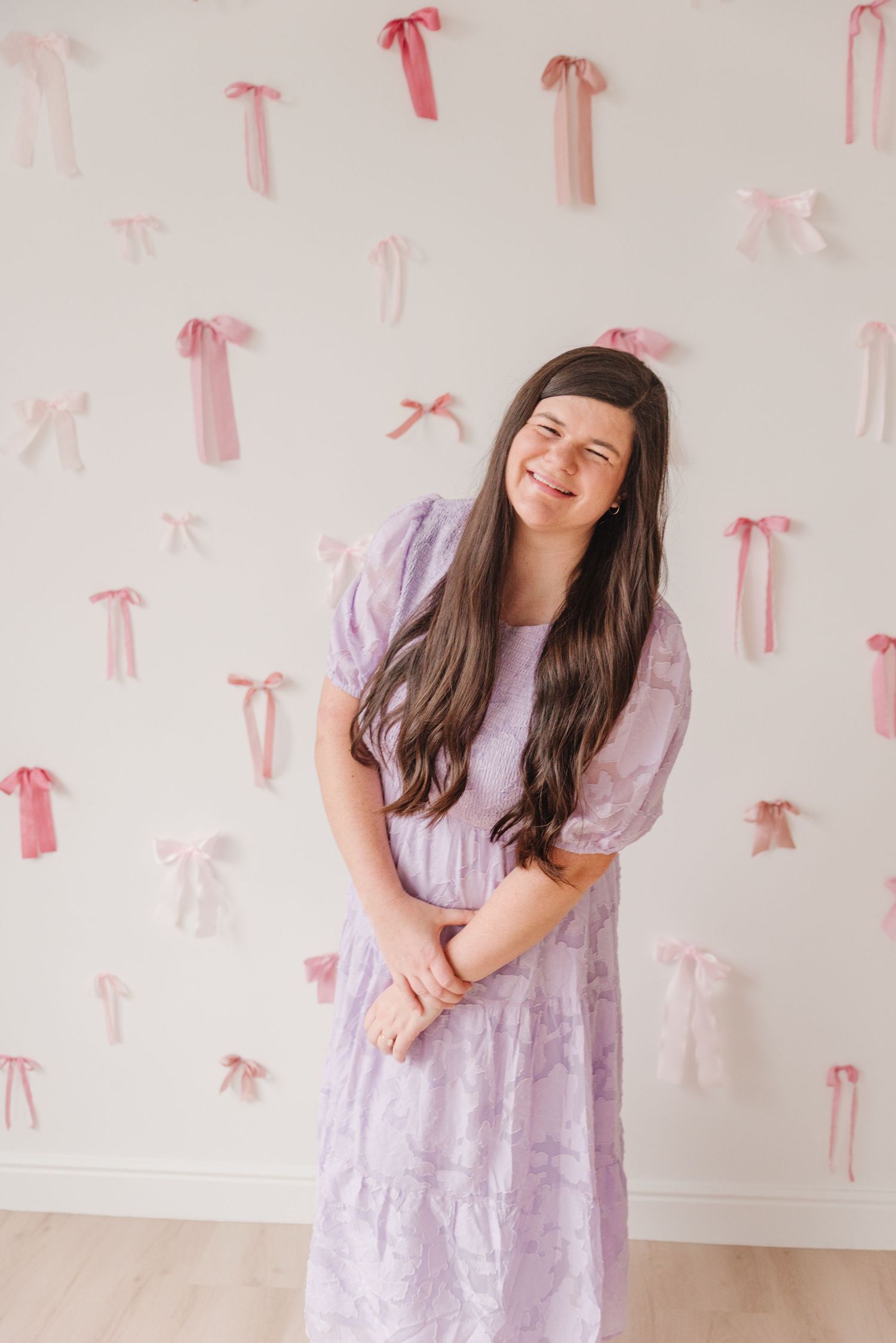 Someone wearing a lavender dress poses against a wall decorated with pink ribbon bows in a cheerful indoor setting.