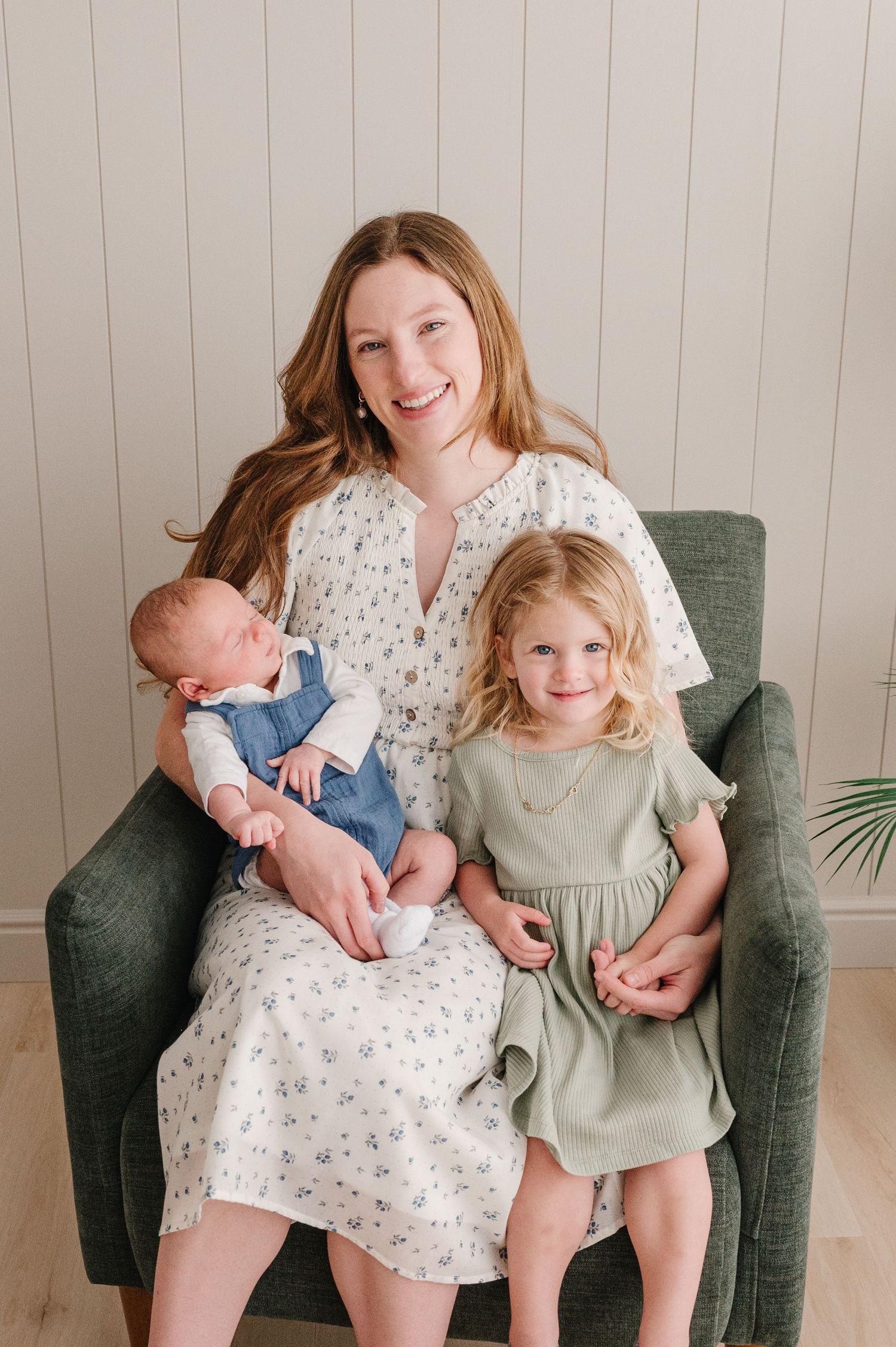 A family sits together on a gray armchair, wearing light floral and sage dresses while holding a sleeping infant.