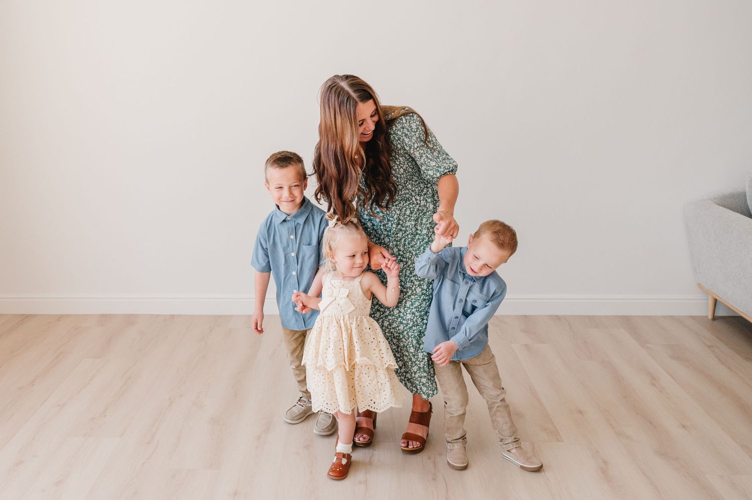 A woman in a floral dress stands with three children on a light wooden floor in a minimalist setting.