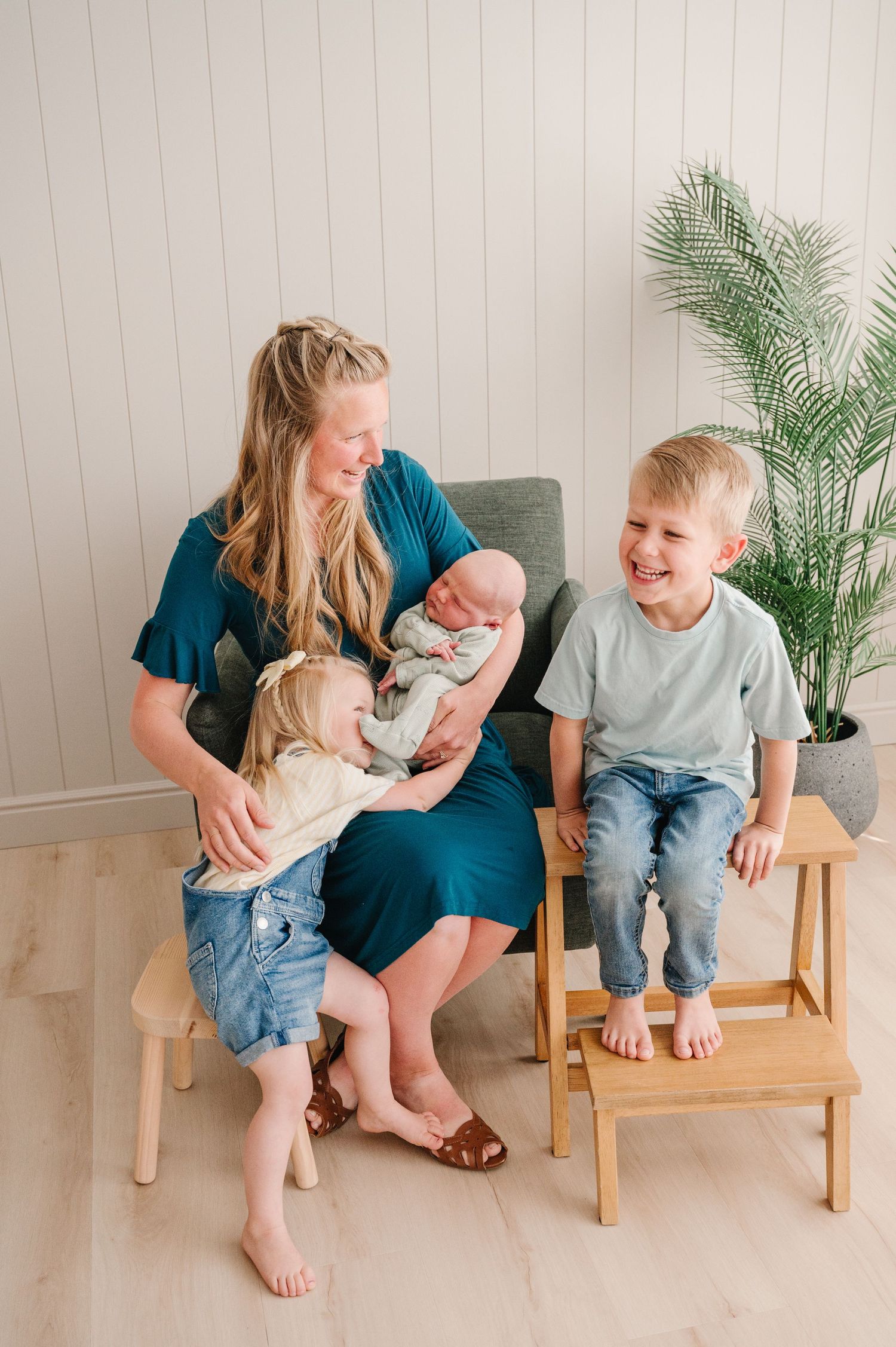 A serene family scene shows tender moments with young children and mother in a minimalist white room with a potted palm.