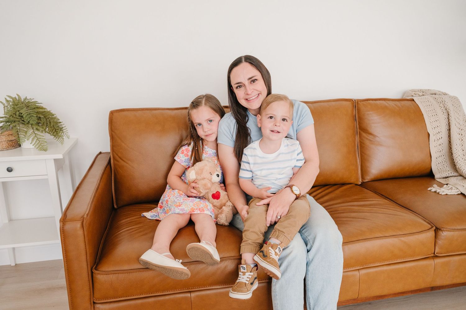 Three people sit together on a brown leather couch in a bright room with a potted plant in the background.