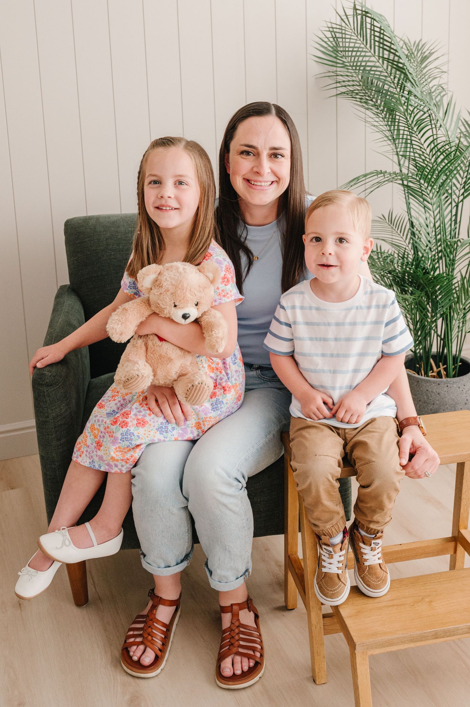 Indoor portrait shows family sitting together on green chair with houseplant, plush teddy bear and hardwood floors.