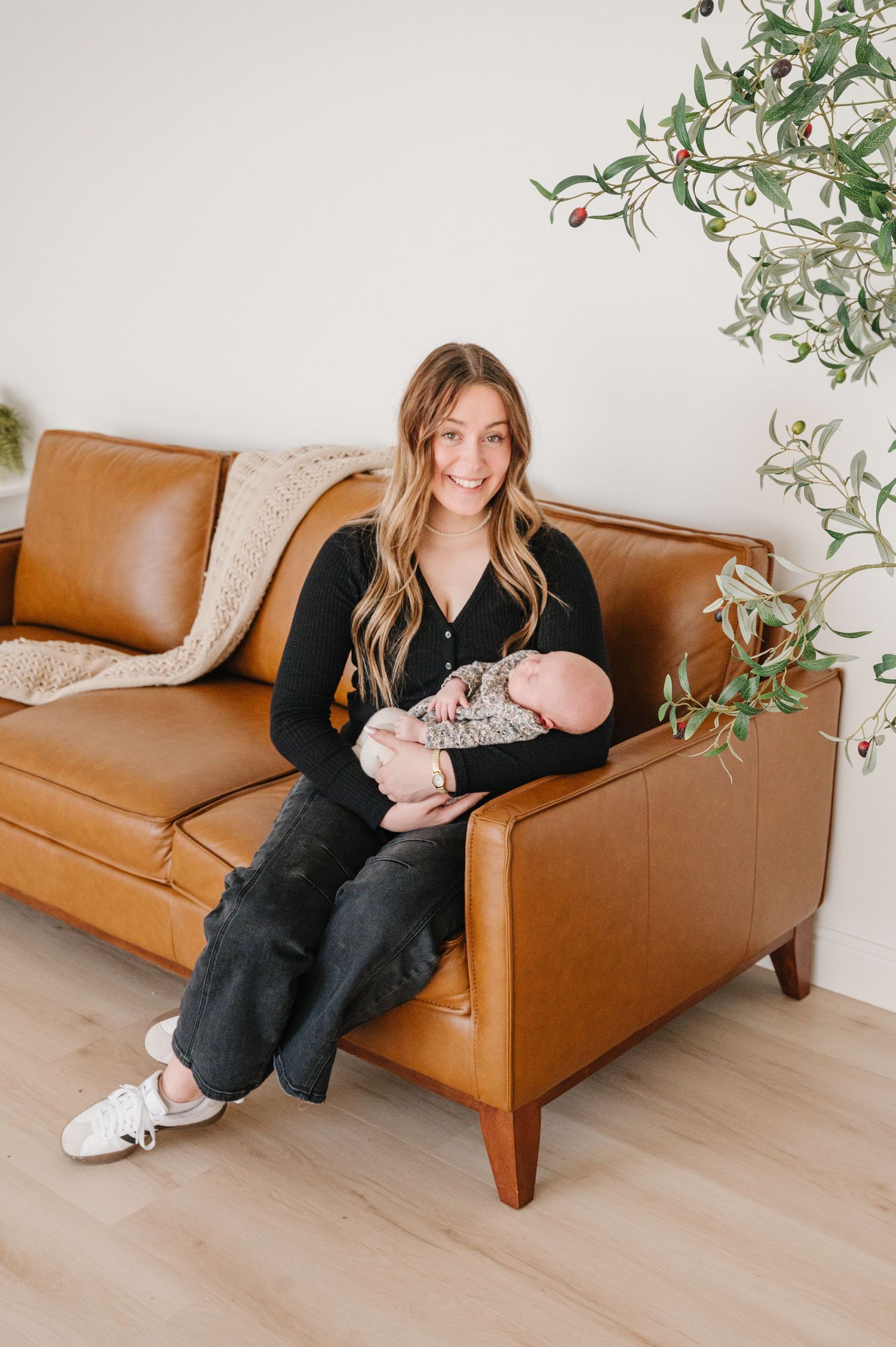 Mother holding sleeping newborn baby relaxes on tan leather couch with hanging plants in minimalist modern living room.