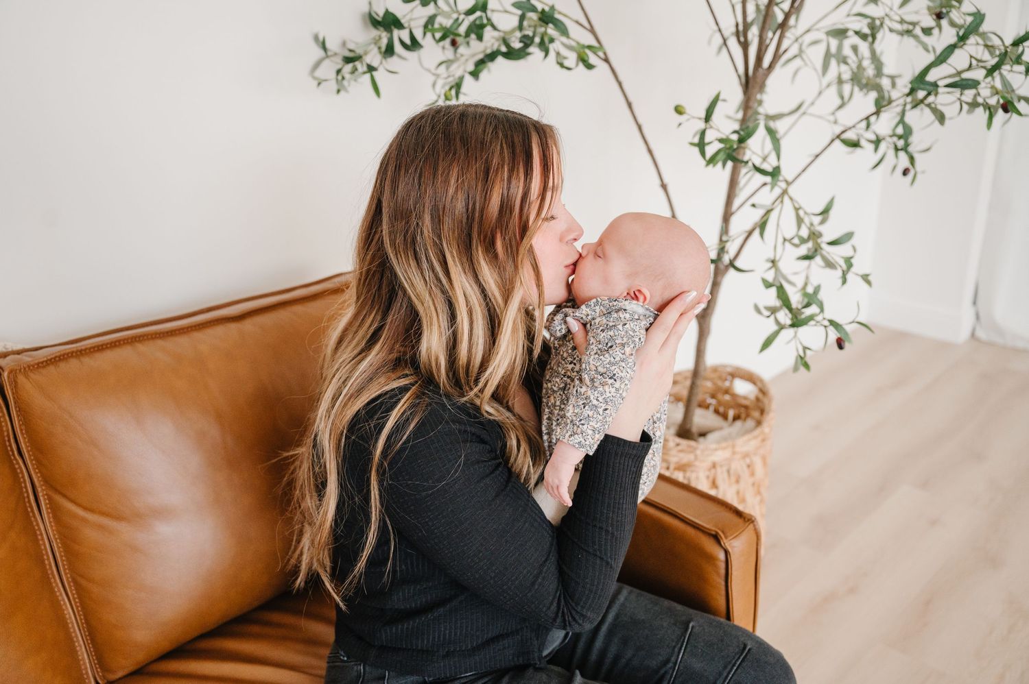 A person in a black sweater holds a baby while sitting on a brown leather couch next to green foliage.