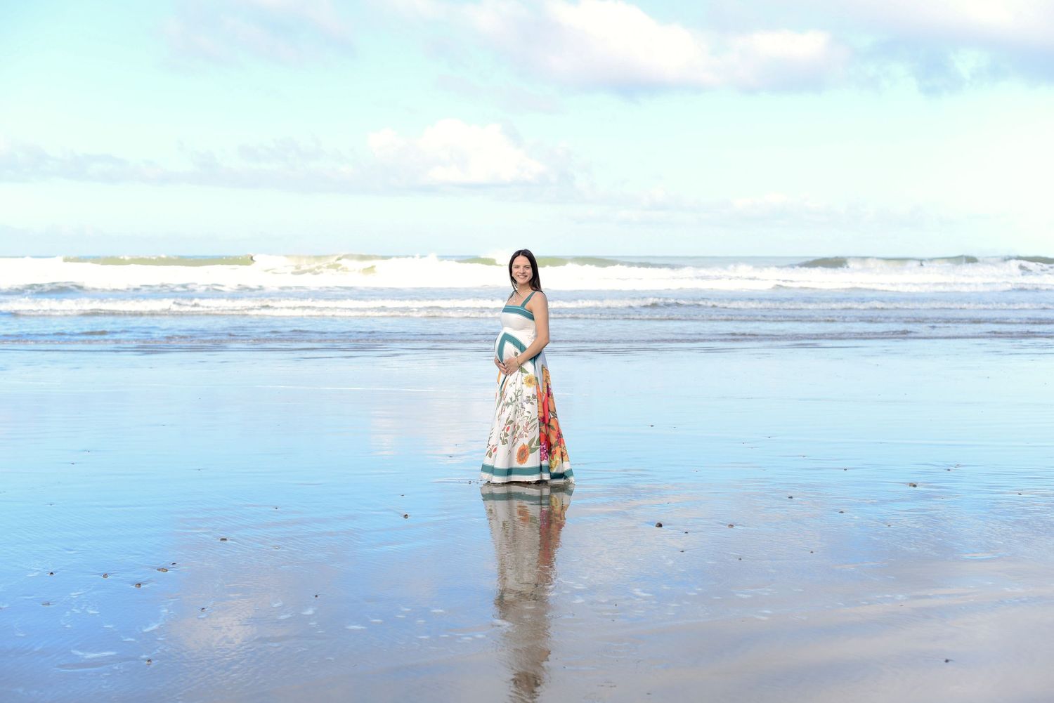 Peaceful maternity portrait on reflective beach with waves and blue sky.