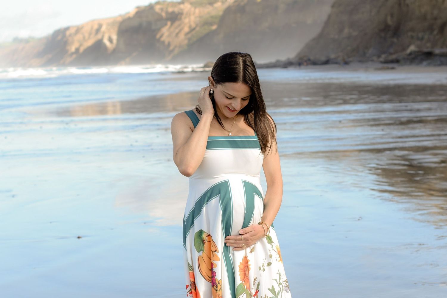 Candid maternity moment on sandy beach with coastal cliffs in background.