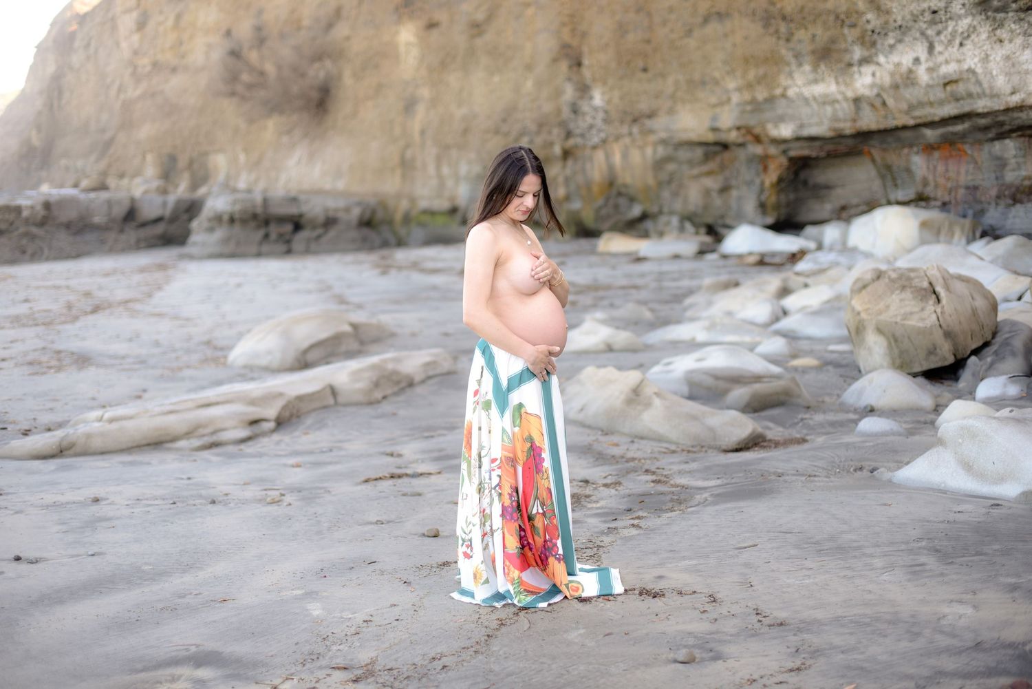Expectant mother in a colorful flowing dress stands contemplatively on a rocky beach.