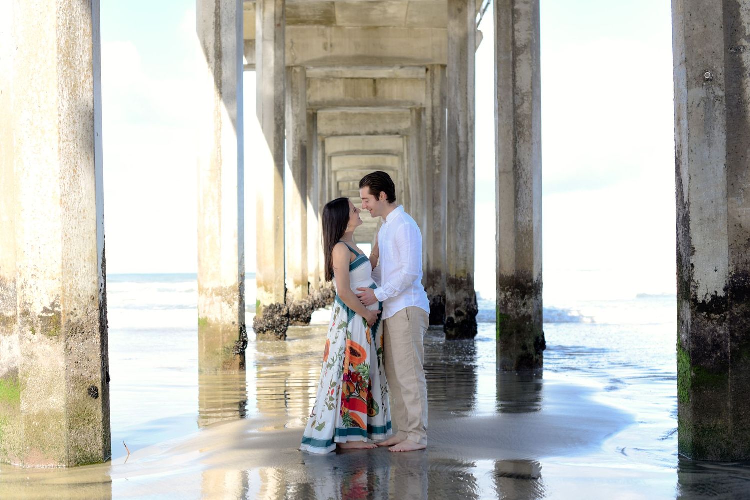 Intimate maternity moment captured beneath pier pillars with ocean backdrop.