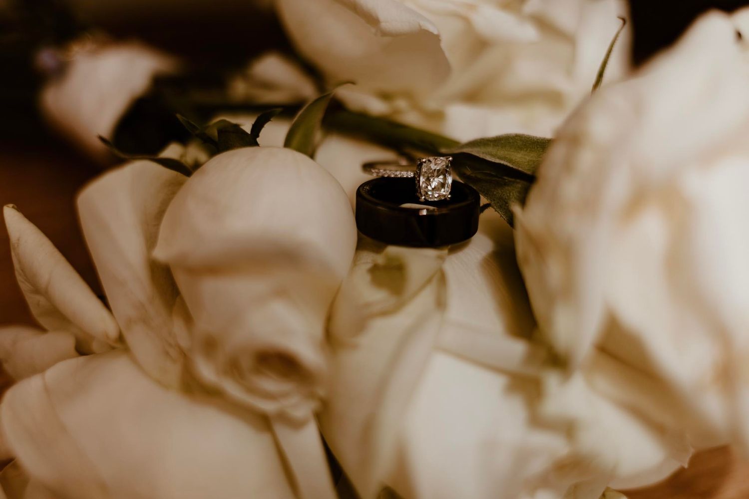 A diamond engagement ring nestles among soft white rose petals in a romantic wedding detail shot.