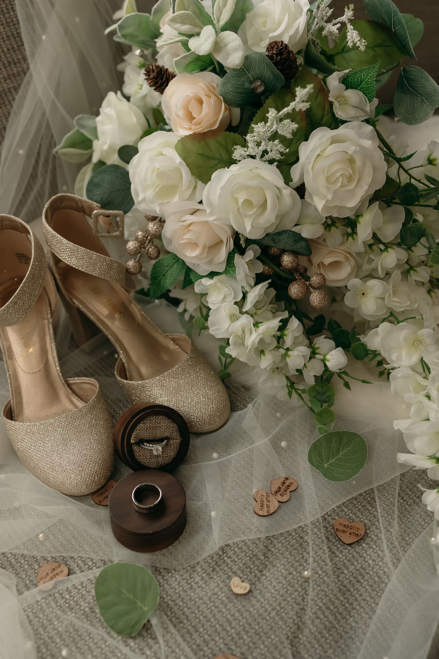 Wedding details flatlay with white and blush rose bouquet, sparkly gold heels, and ring box on tulle fabric.