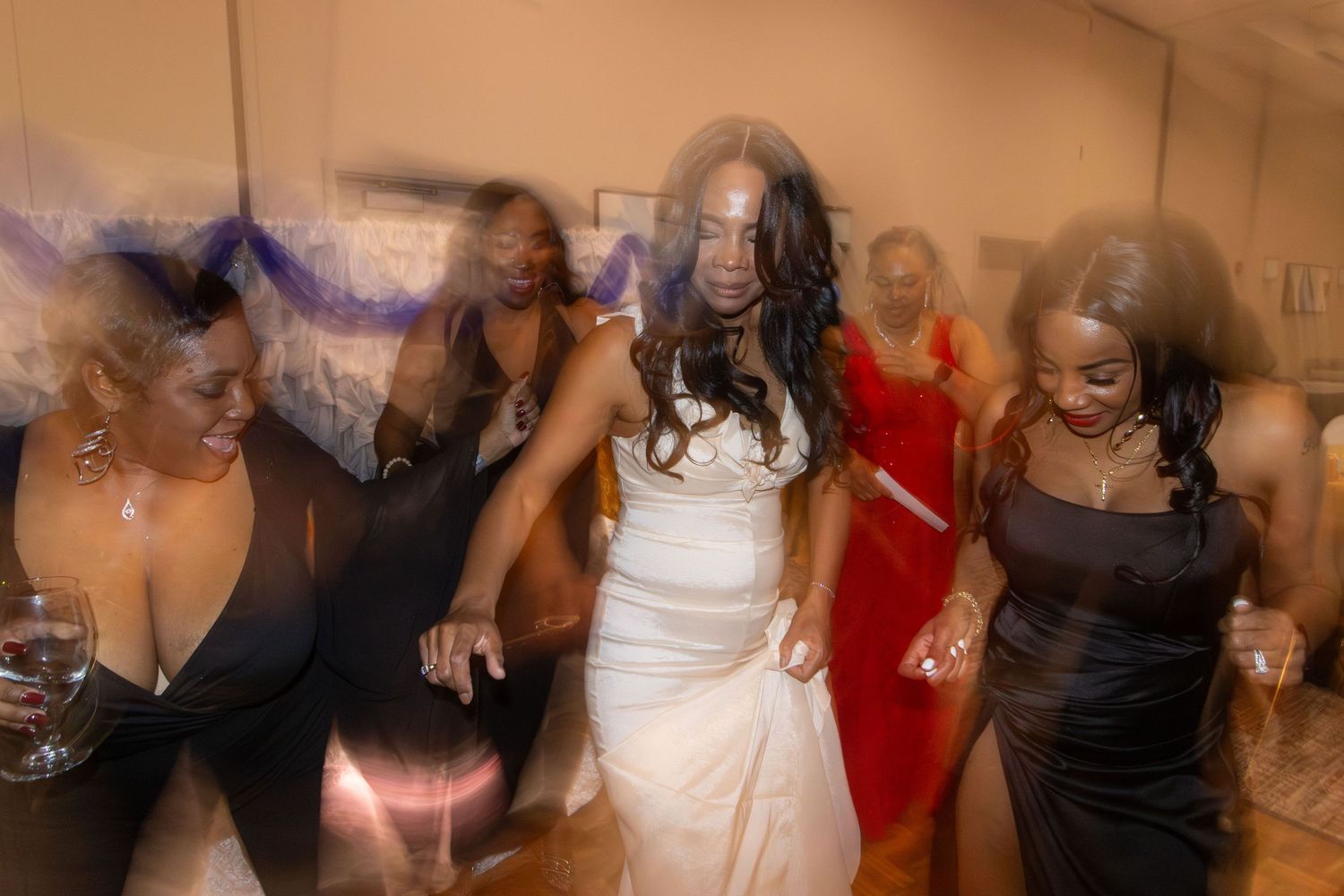 Motion blur captures people dancing and celebrating with someone in a white dress at what appears to be a wedding reception.