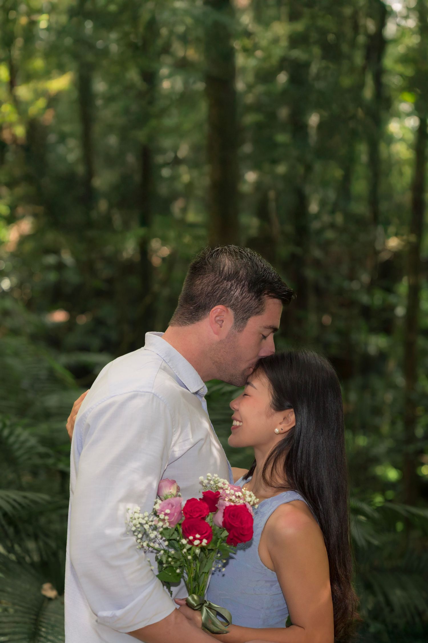 Romantic couple sharing a tender moment in a lush forest setting with red roses and baby's breath bouquet.