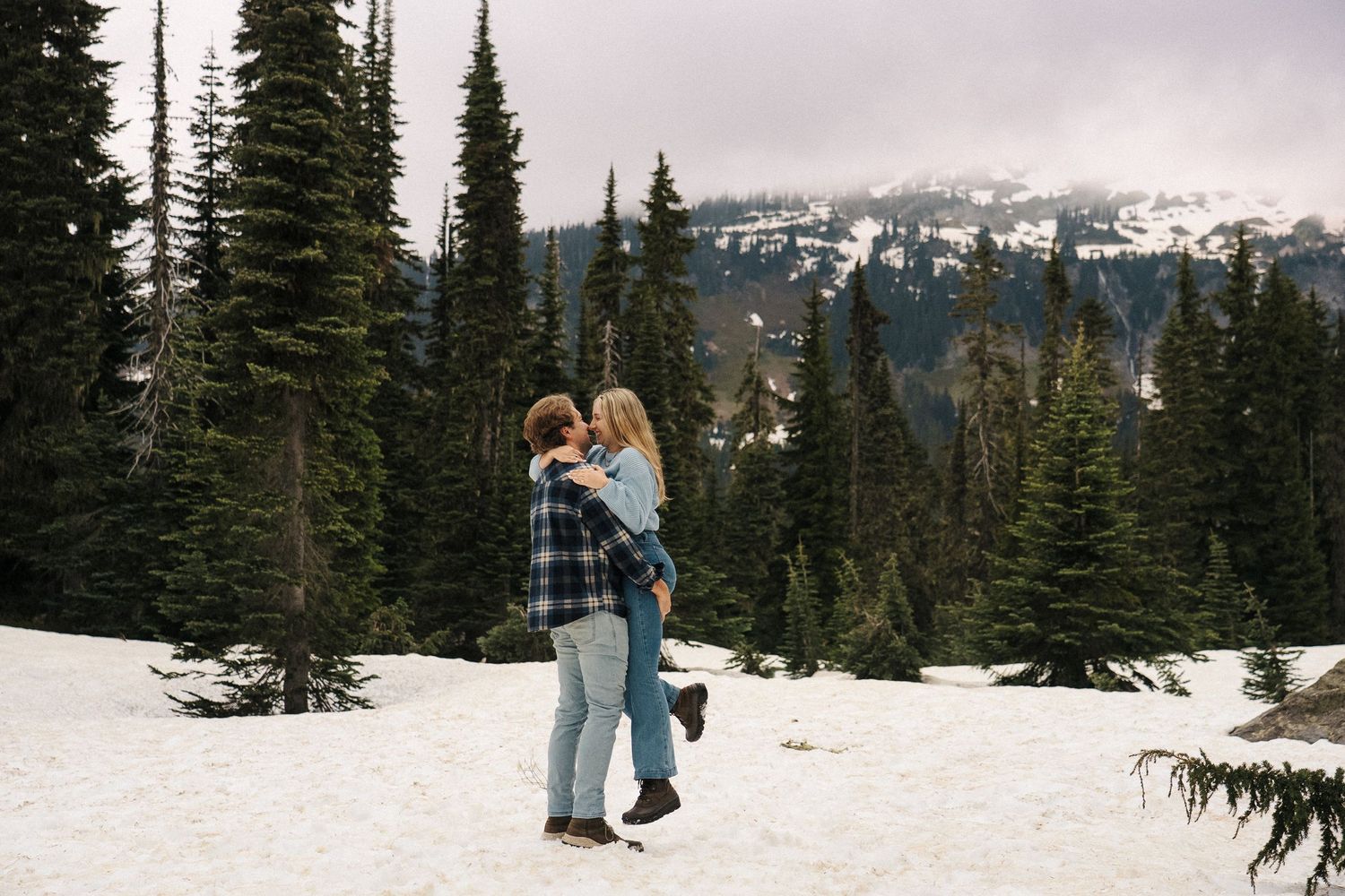 A couple embrace in a snowy alpine landscape surrounded by tall evergreen trees and mountain peaks in the distance.