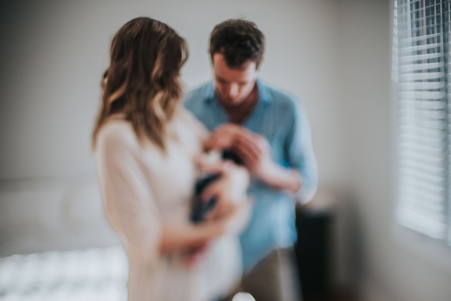 Two people standing in front of a window with soft blurred focus caring for a newborn baby together indoors.