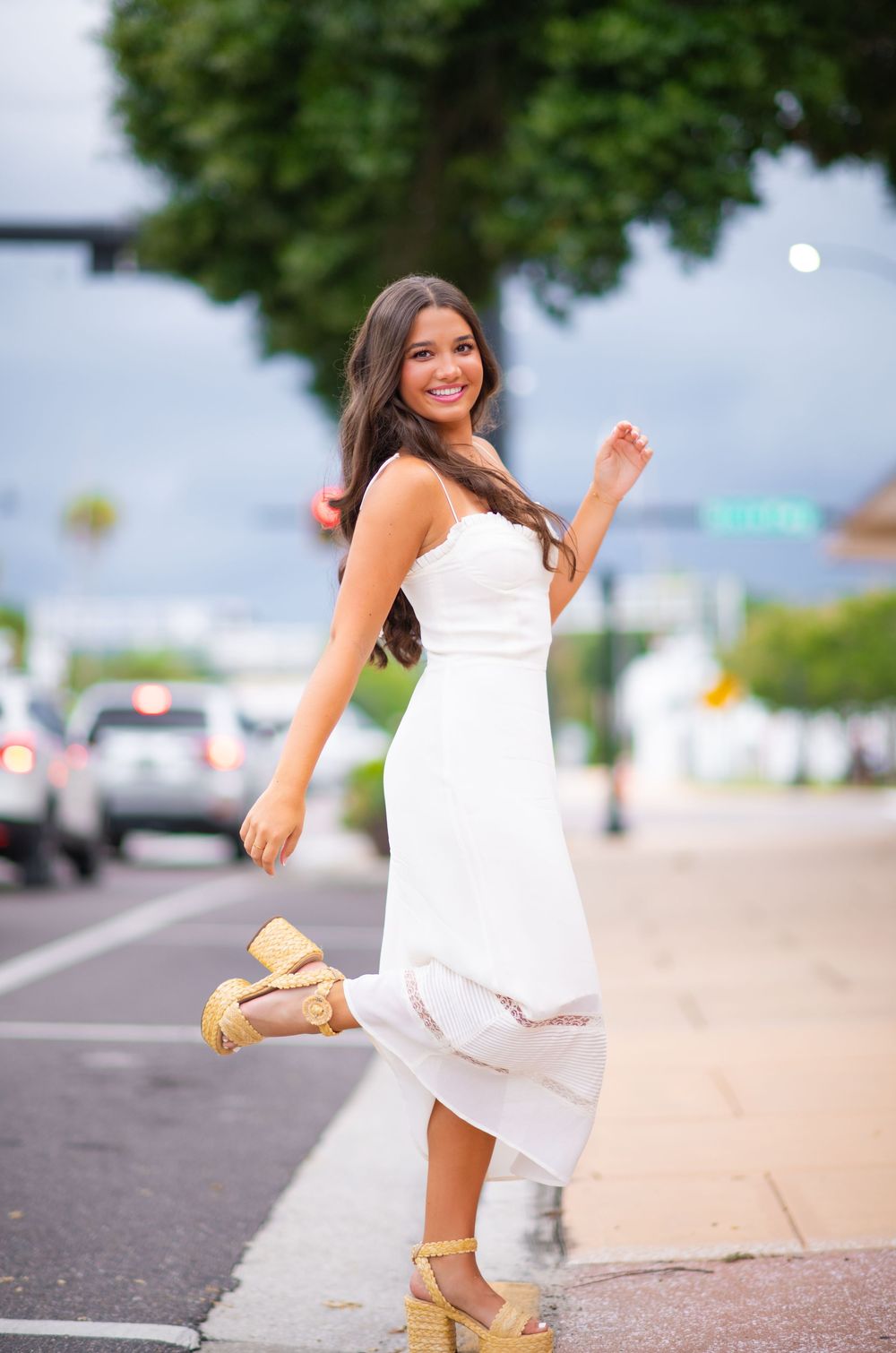 Young woman in white sundress and platform sandals twirls playfully on a city street.