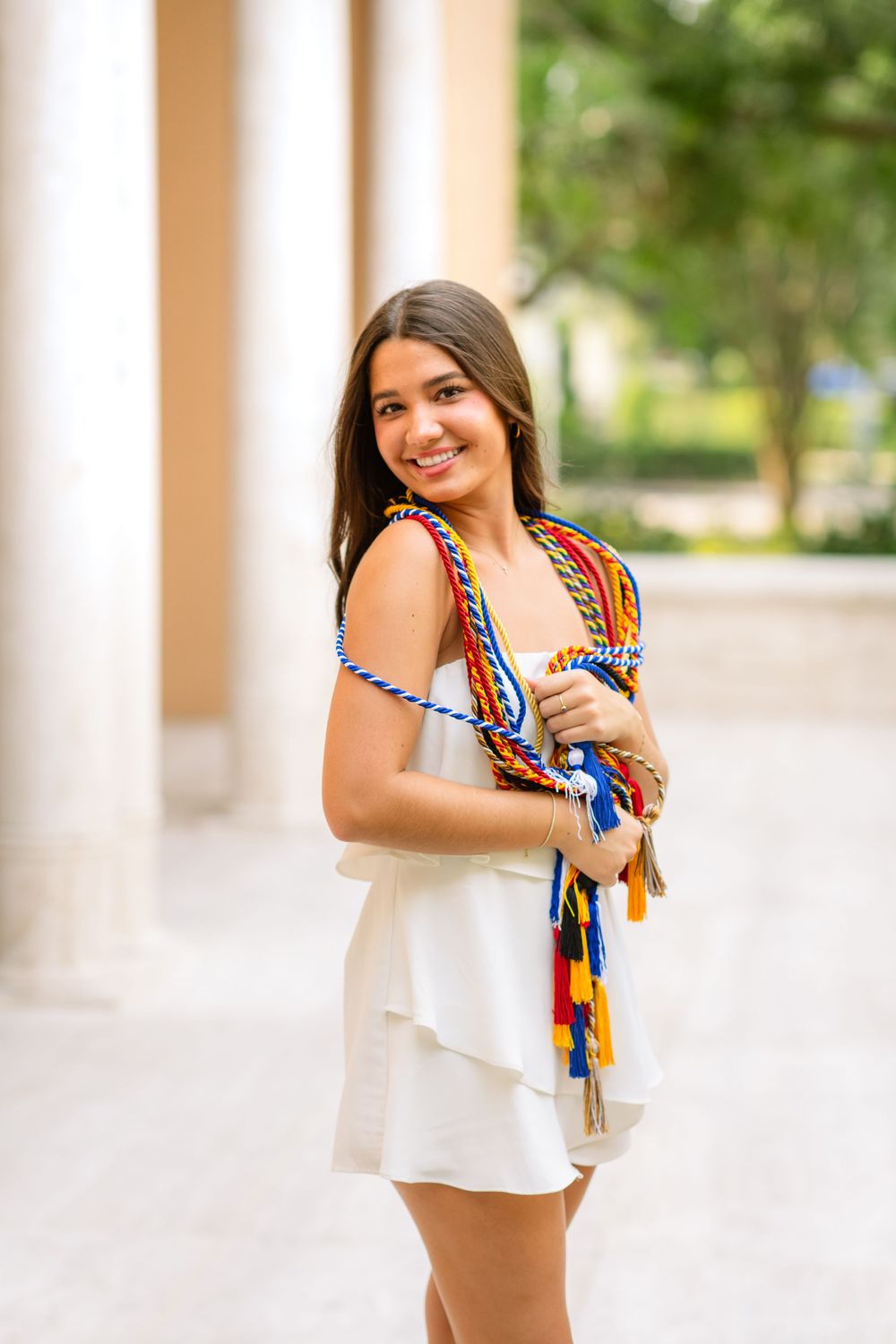 A woman in a white outfit with colorful patterned scarf stands between outdoor columns.