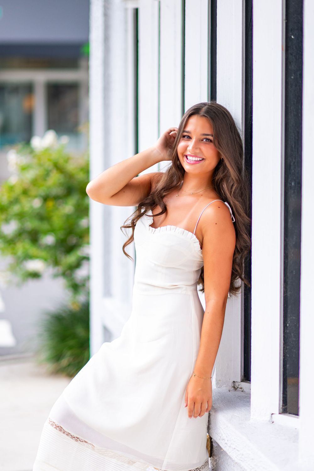 A person in a white dress sits casually against a window frame in a summery outdoor setting.