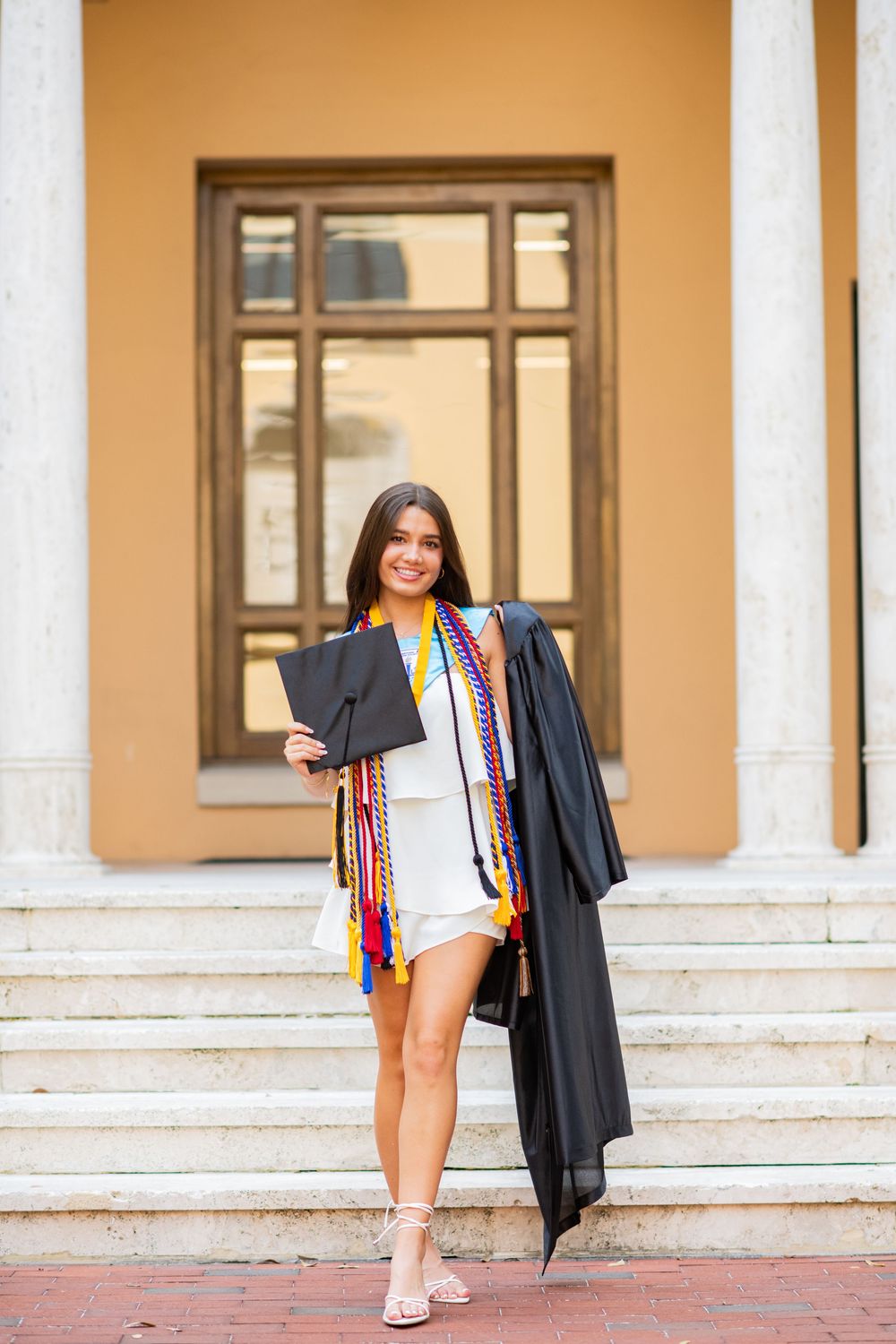 A graduate poses on stone steps with cap, gown and colorful graduation cords.
