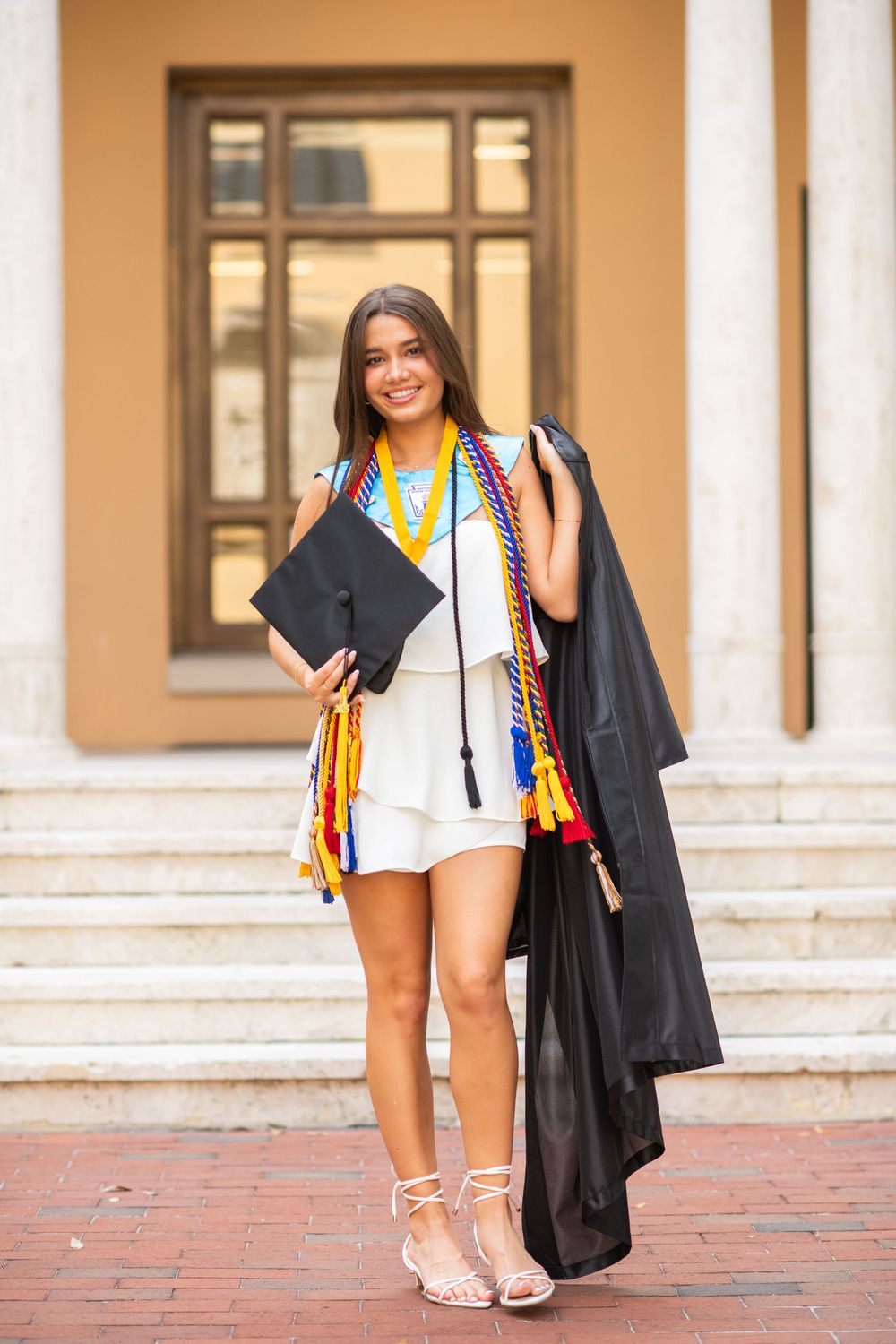 Recent graduate holds cap while wearing graduation stole and white outfit on campus steps.