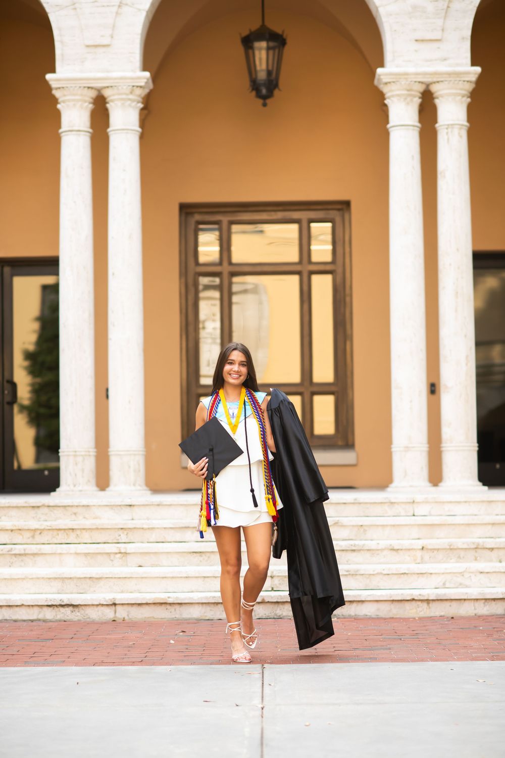 Graduate holds diploma while standing on steps of building with classical architecture and arches.