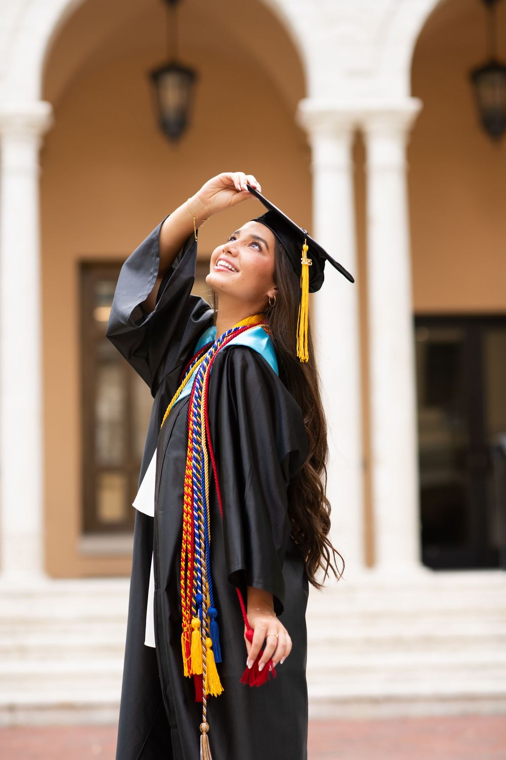 Graduate adjusts graduation cap tassel while smiling under ornate archways of campus building.