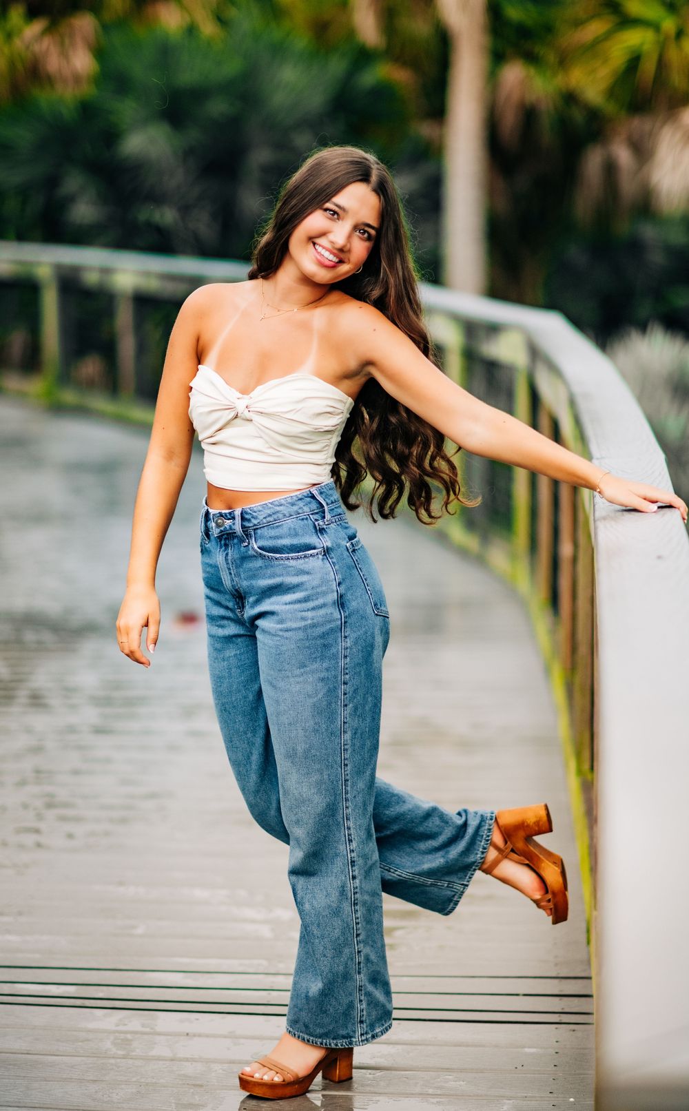 A woman in a white crop top and wide-leg jeans poses playfully on a wooden bridge.