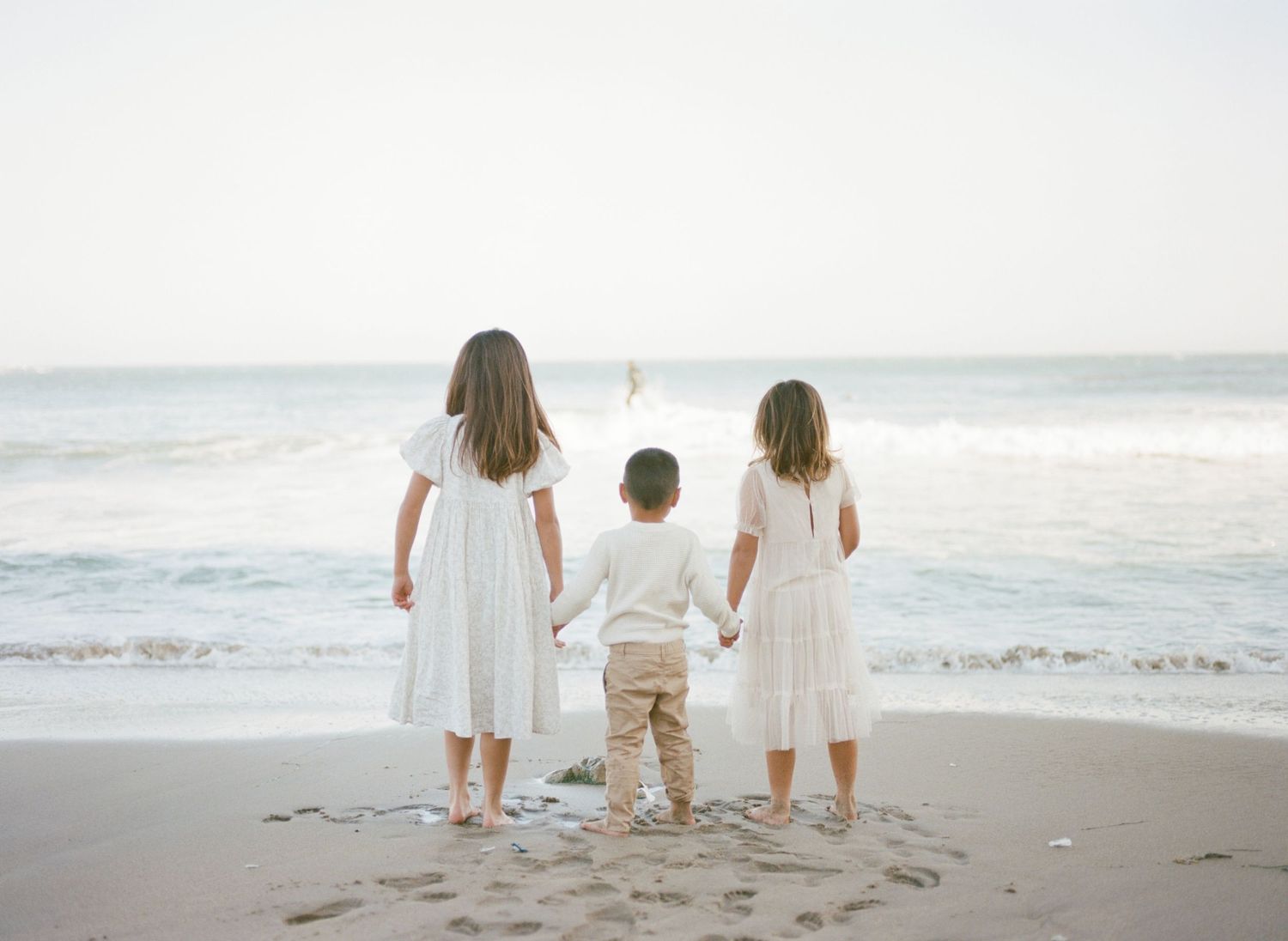 A family wearing white clothing walks together along a sandy beach at sunset.