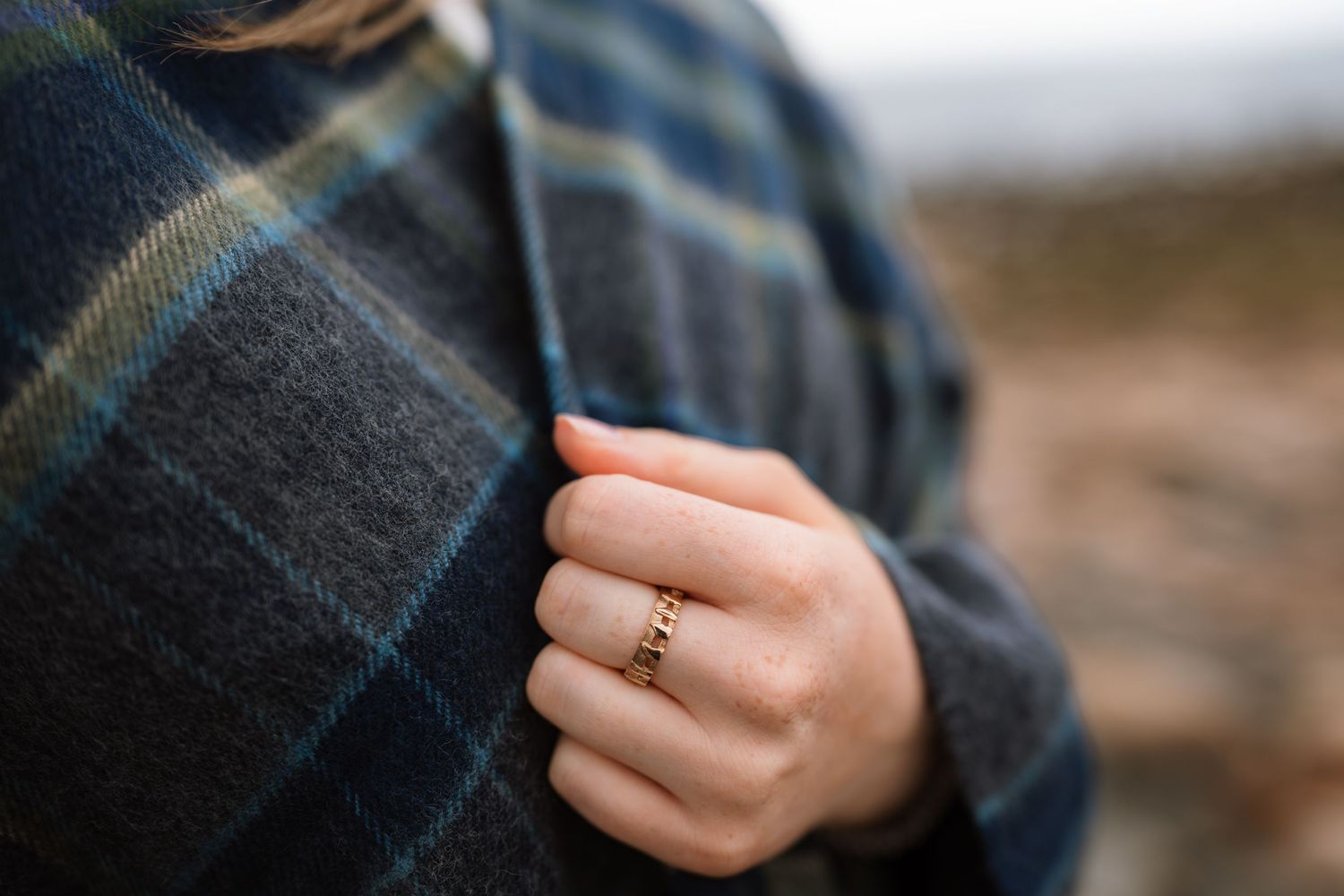 A delicate diamond ring adorns a hand against a dark plaid fabric background.
