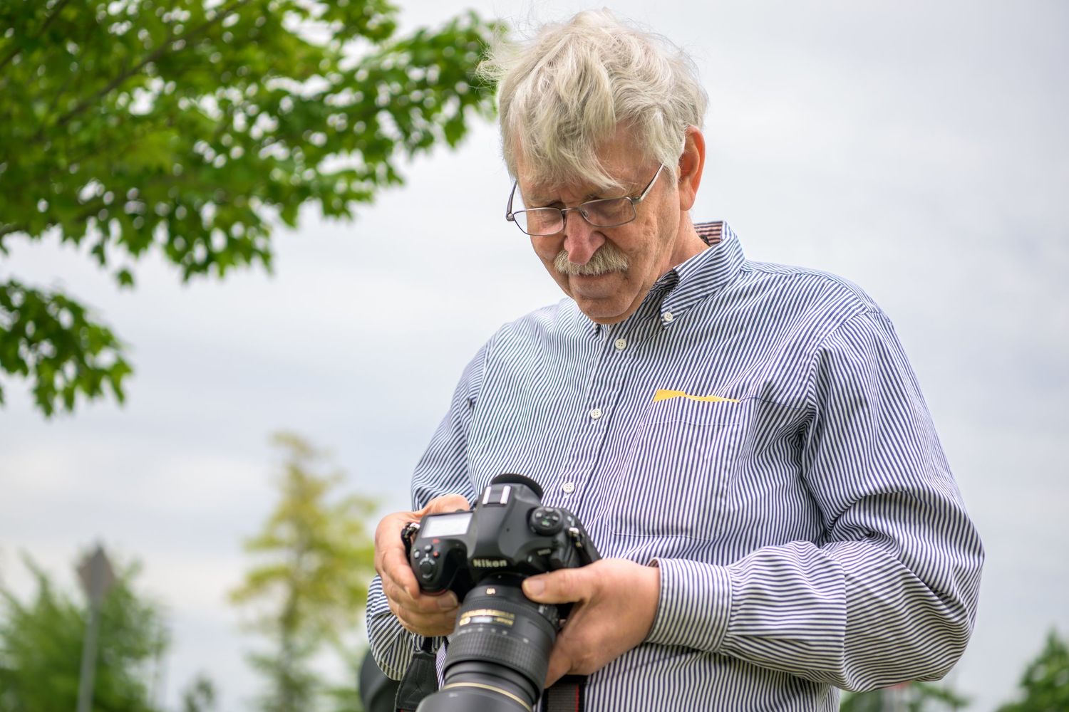 A person wearing a light blue button-down shirt reviews photos on a DSLR camera outdoors with green trees in the background.