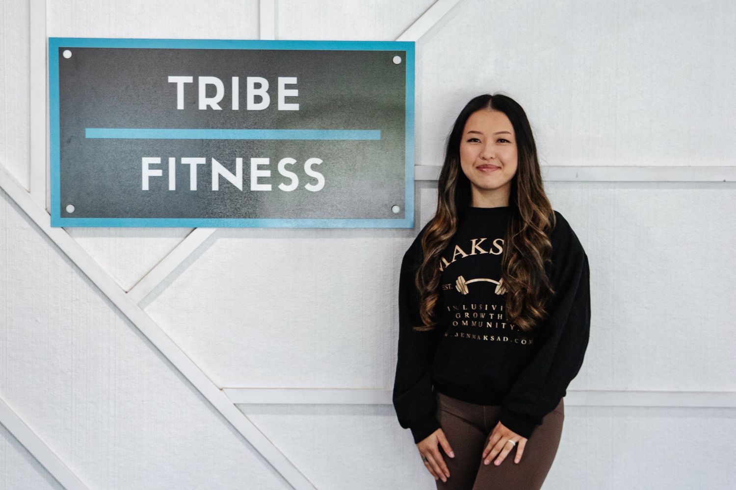 Woman smiling in front of a wall sign that reads "Tribe Fitness," wearing a sweatshirt and standing in a casual pose
