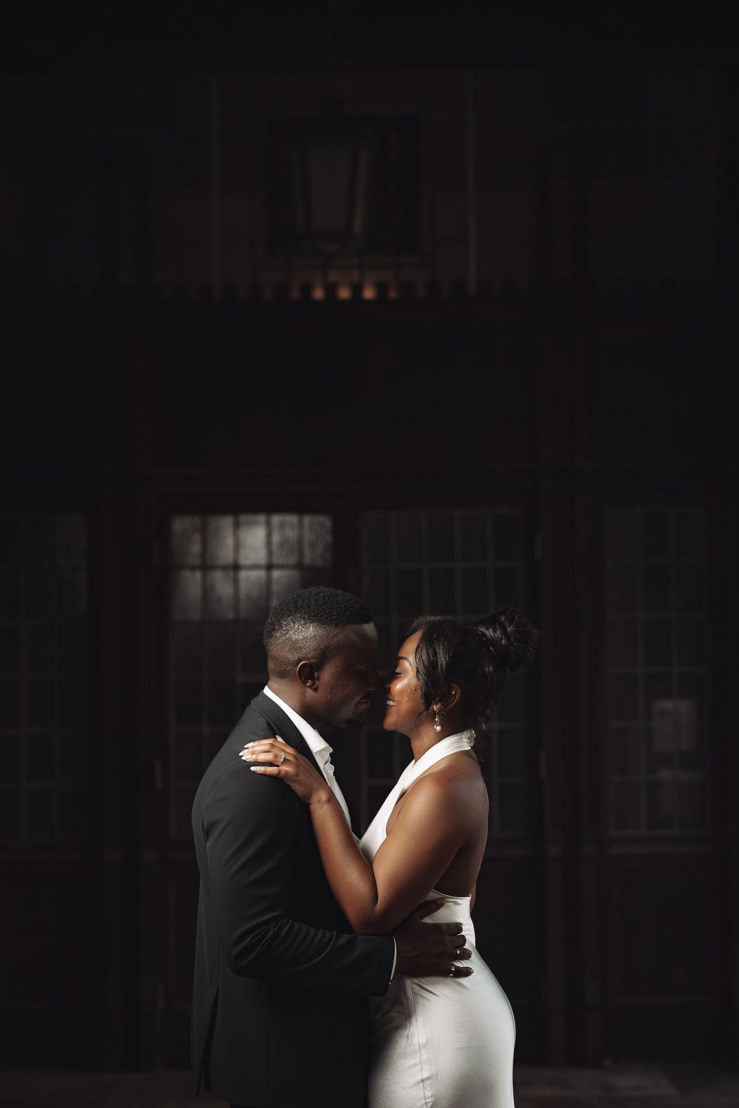 A couple shares an intimate moment in formal wedding attire against a dark dramatic background.