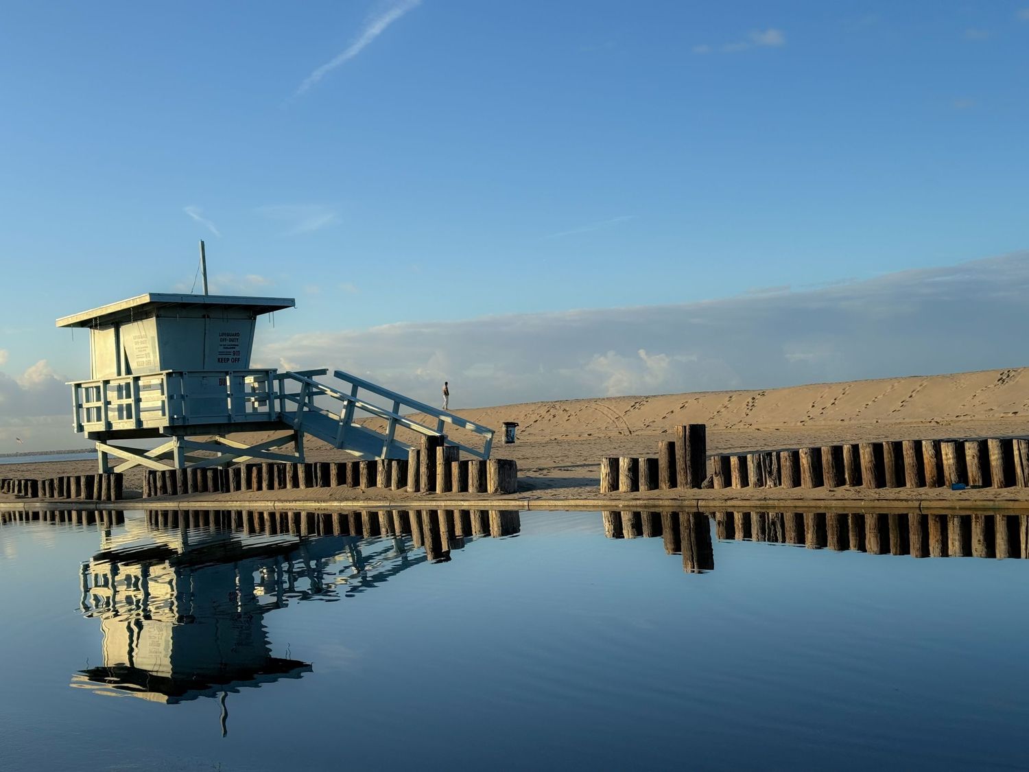 Lifeguard tower and wooden pier reflecting perfectly in calm ocean water during a bright sunny day.
