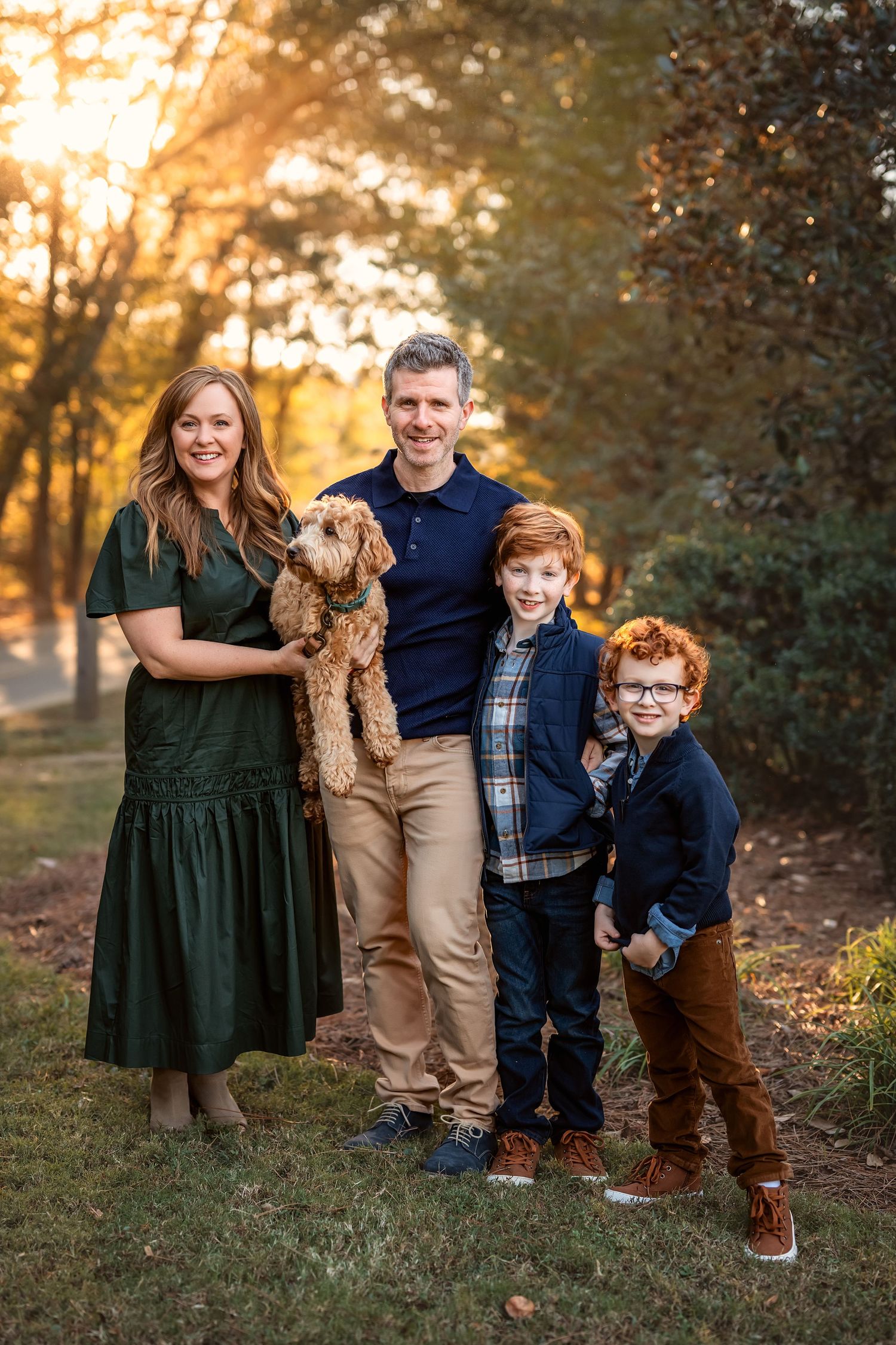 A family with two children and their golden doodle dog pose together on a grassy hill during golden hour sunset.