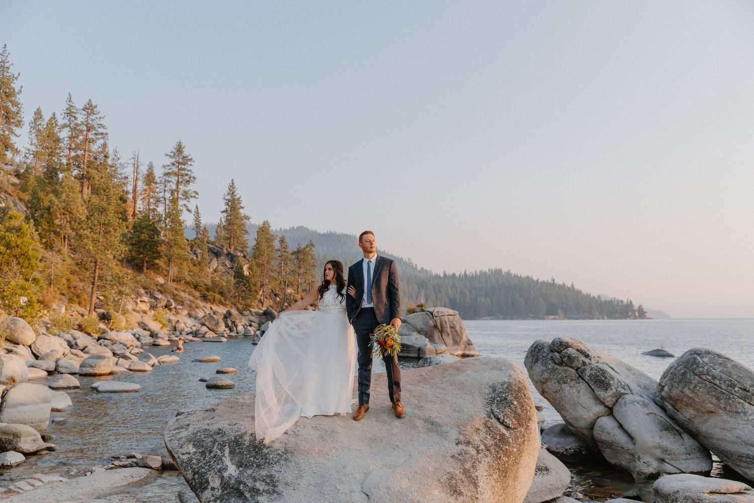 A couple stands on large boulders along a scenic lake shore with pine trees and mountains in the background at sunset.