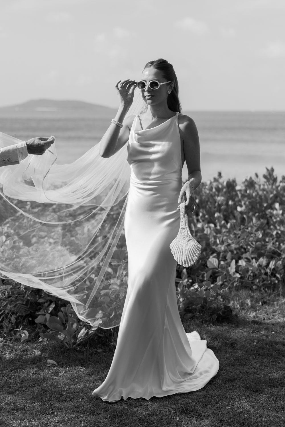 A flowing white wedding dress veil billows gracefully in the wind against a scenic coastal landscape background.