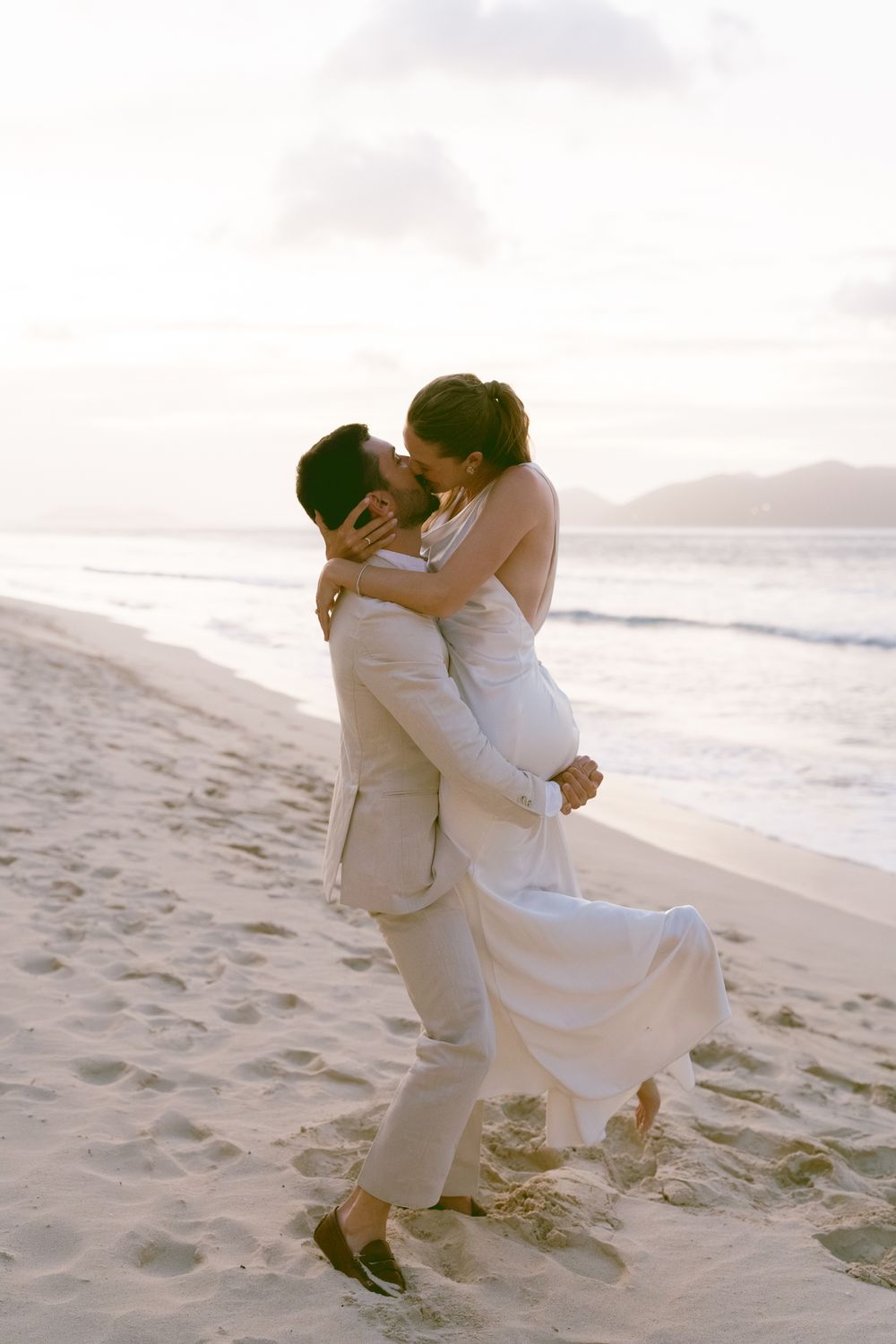 A romantic couple embraces on a sandy beach at sunset, with gentle waves in the background.