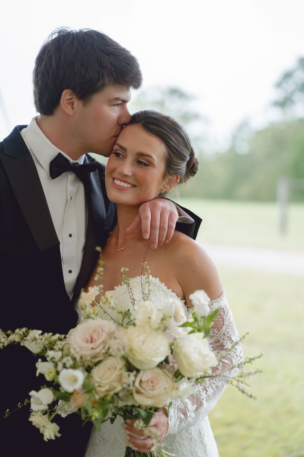 Groom in black tuxedo kissing bride on cheek as she smiles holding white and blush rose bouquet outdoors.