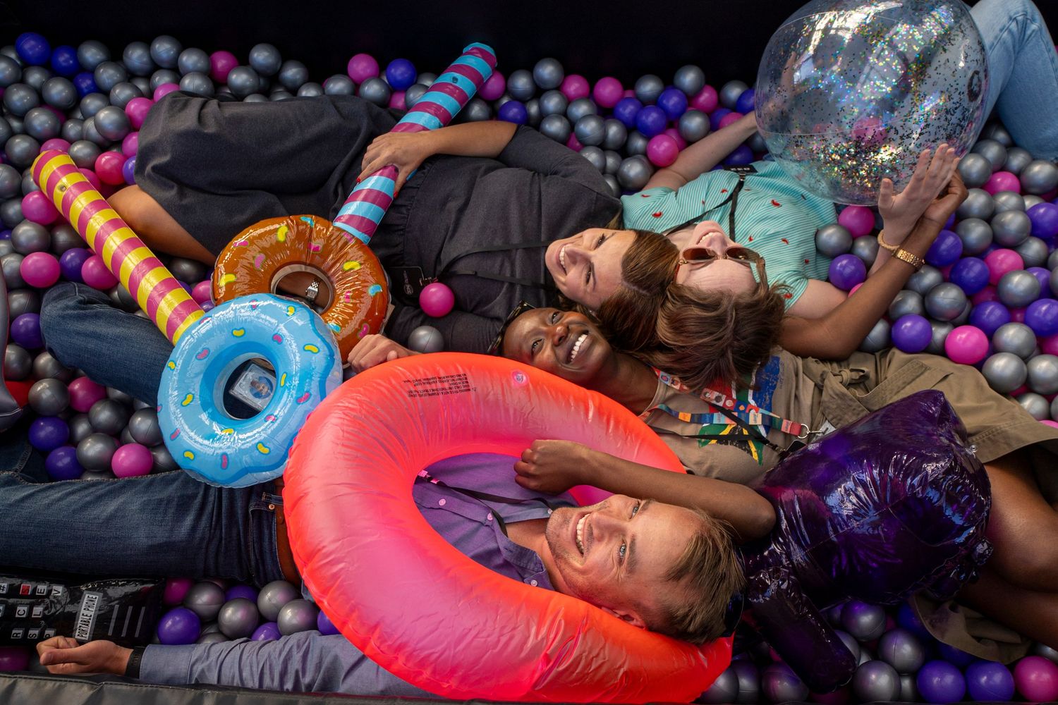 People lying playfully on colorful inflatable pool toys surrounded by plastic balls.
