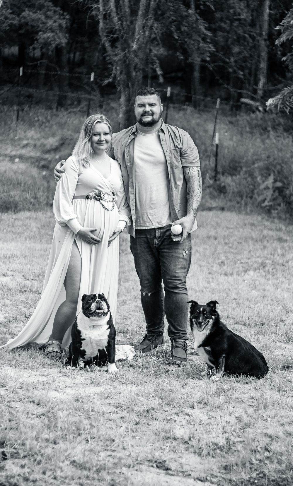 Black and white outdoor photo shoot with couple and their black dog sitting together in a grassy field.