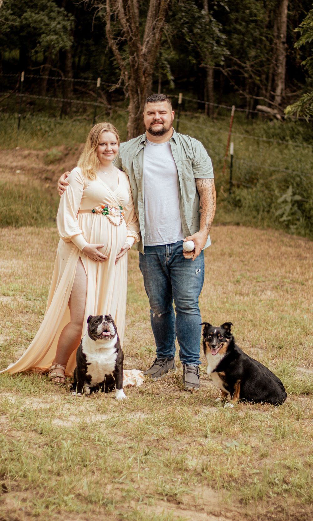 A couple in casual attire stands outdoors with their two small dogs on a grassy field during sunset.