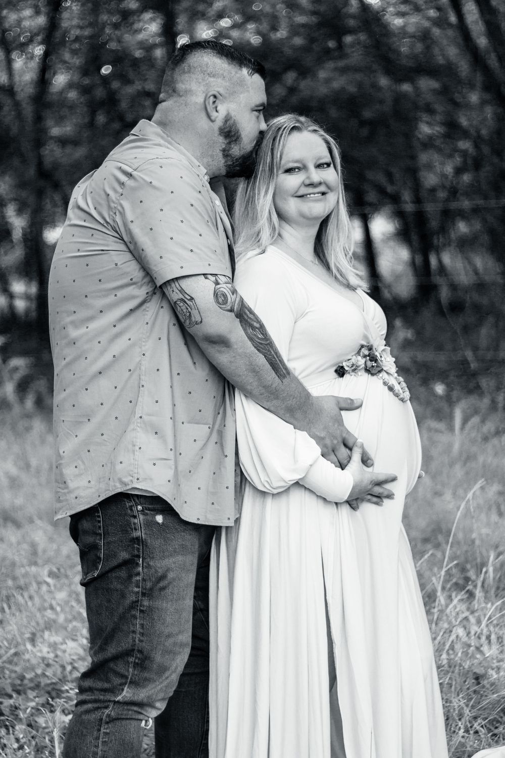 A black and white outdoor maternity photo showing a couple embracing in a peaceful woodland setting.