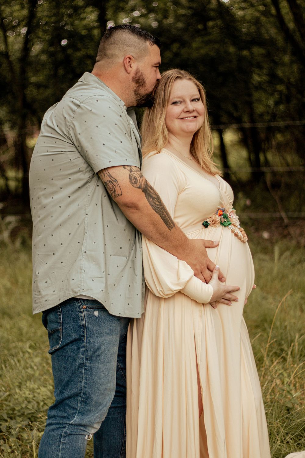 A couple embraces in a natural outdoor setting, with her wearing a flowing cream dress and him in a casual button-up shirt.