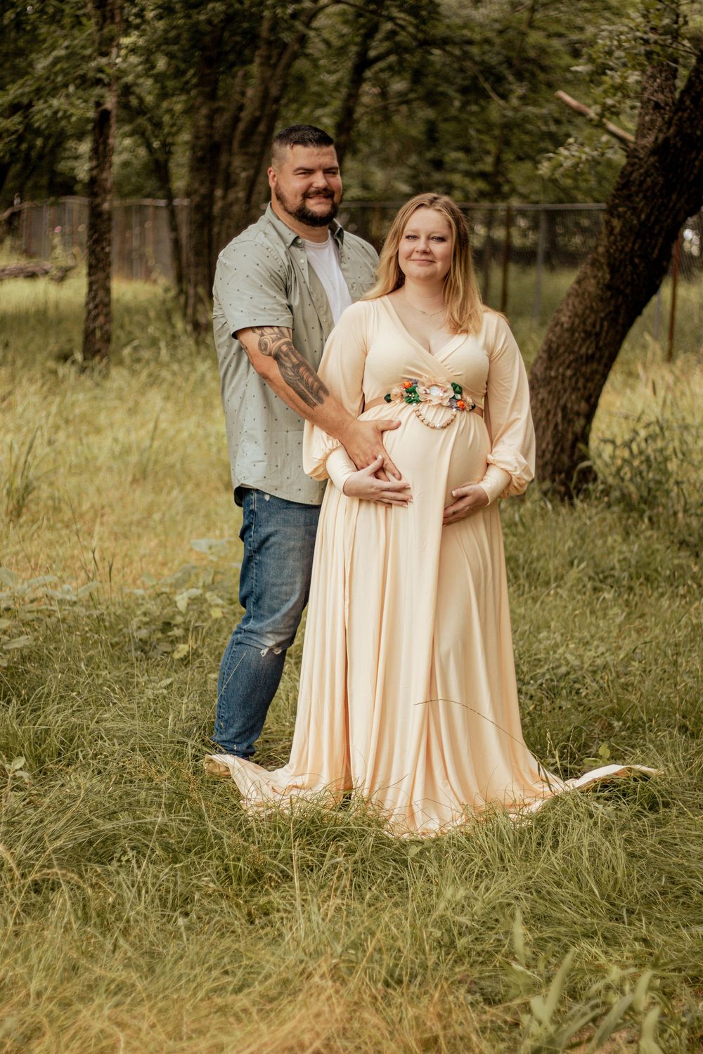 A couple poses together in a natural outdoor setting with trees, with the woman wearing a flowing cream-colored dress.