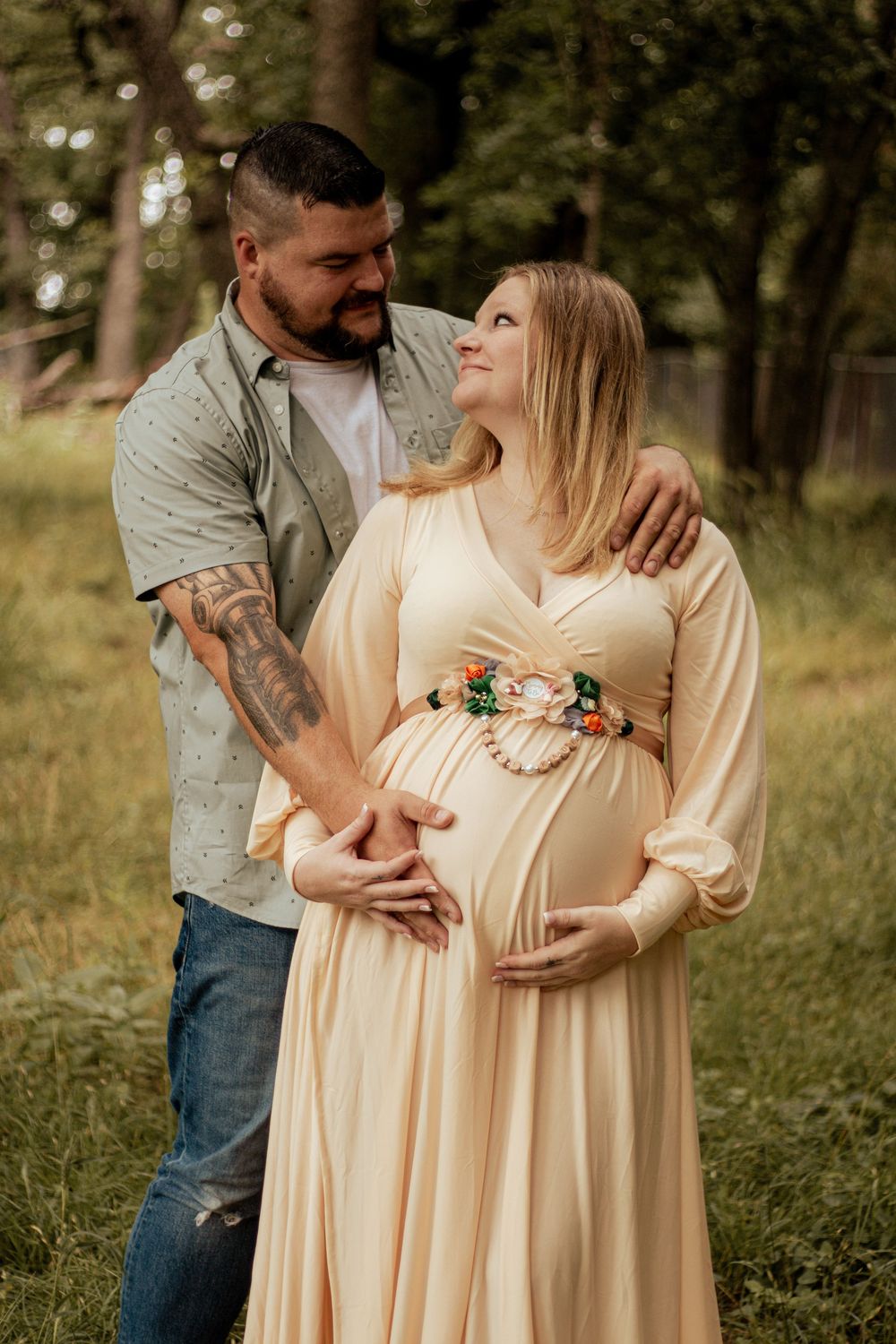 Pregnant woman in cream dress with floral sash embraces partner in woodland maternity photo shoot setting.