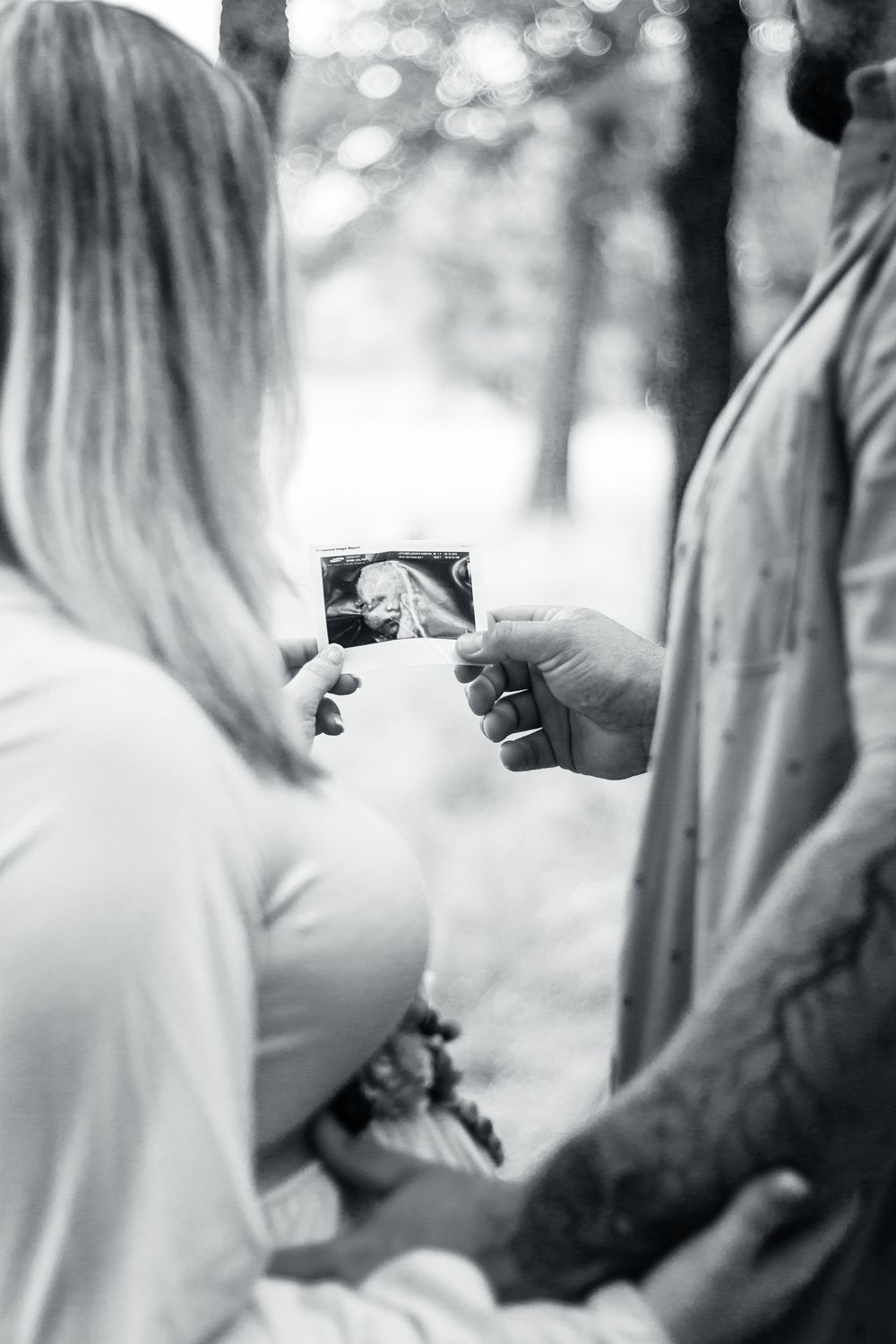 Black and white image shows two people looking at a small ultrasound photo together outdoors in a natural setting.